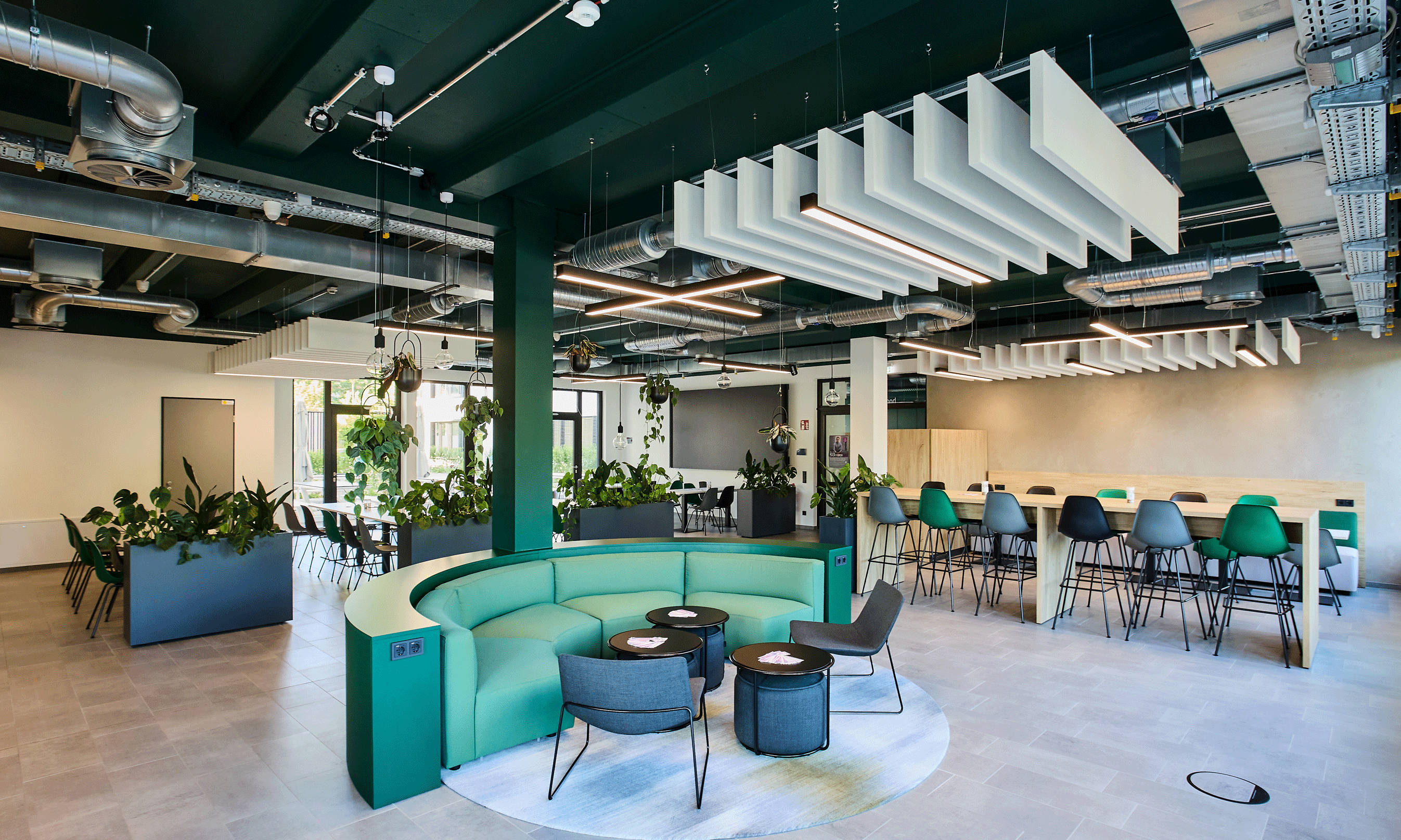 Modern office space with green sofa corner, conference tables and lots of plants, open ceiling with ventilation elements.