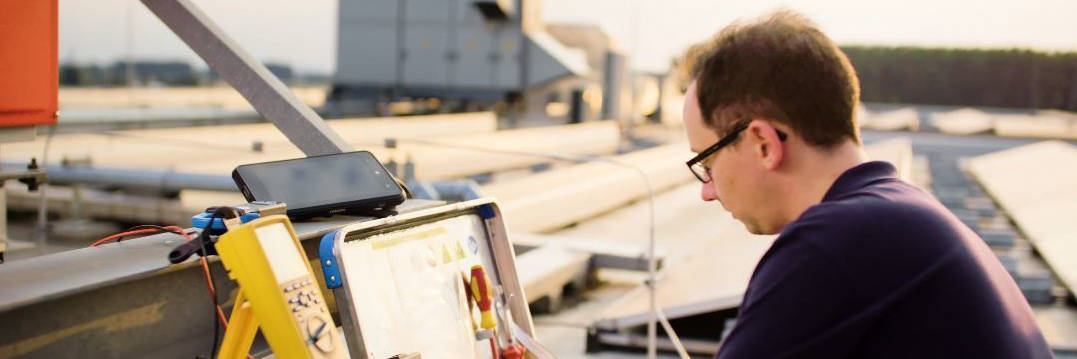 Specialist carrying out technical maintenance on a flat roof, with a multimeter and control box.