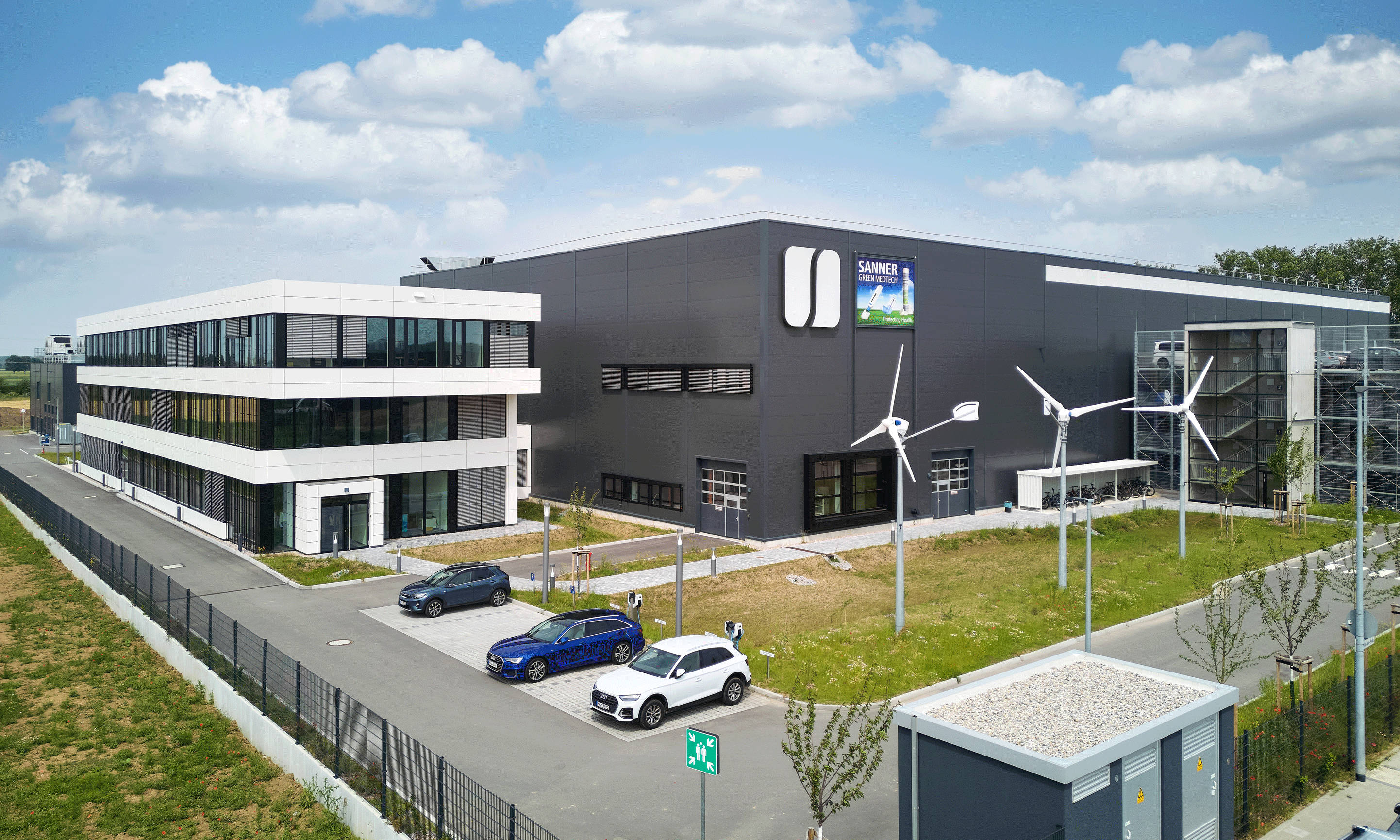 Modernes Büro- und Industriegebäude mit Windturbinen im Vordergrund, blauer Himmel im Hintergrund.