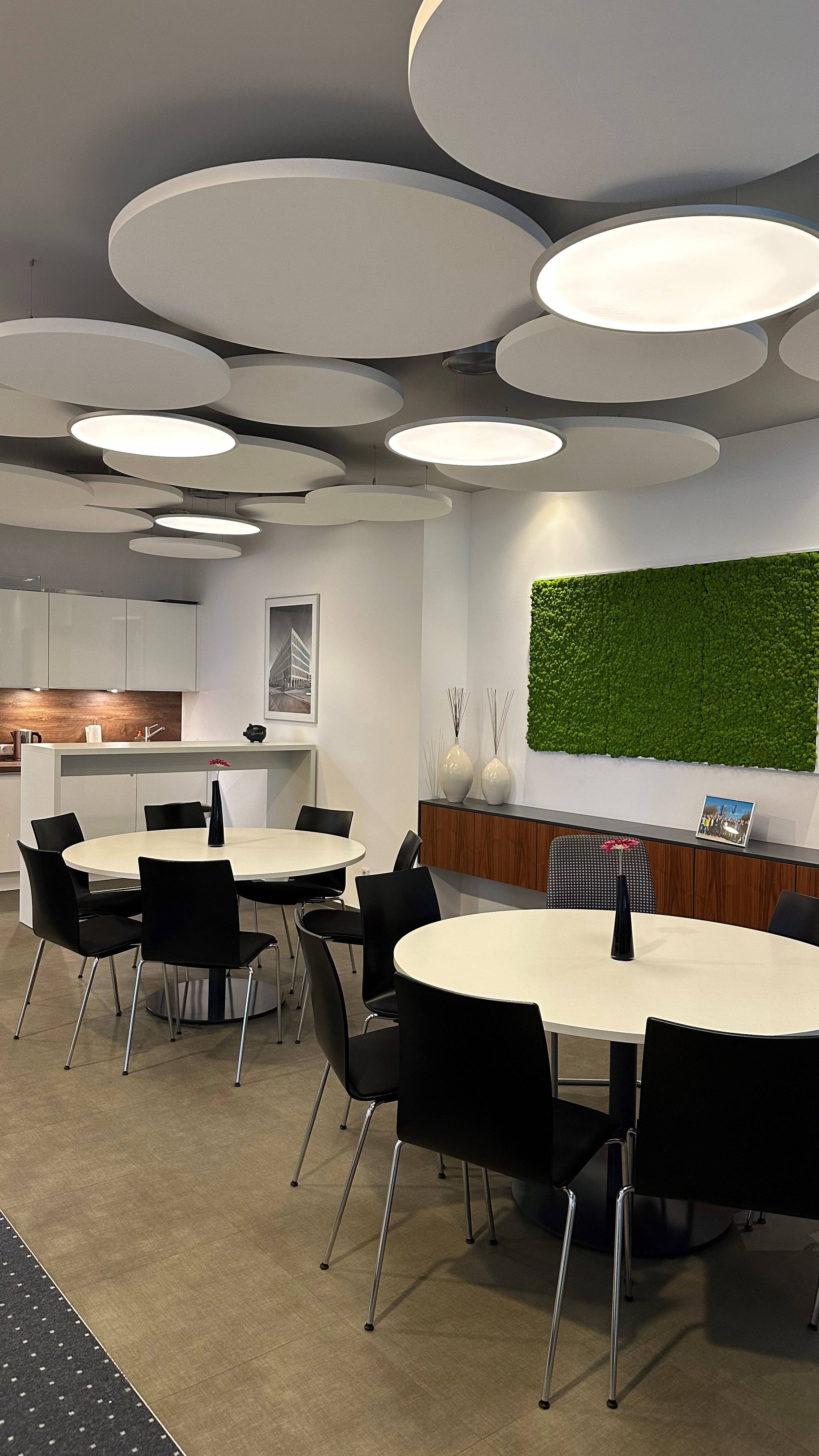 Moderner Büro-Pausenraum mit runden Tischen, schwarzen Stühlen und innovativer Deckenbeleuchtung. Wanddekoration aus grünem Moos.