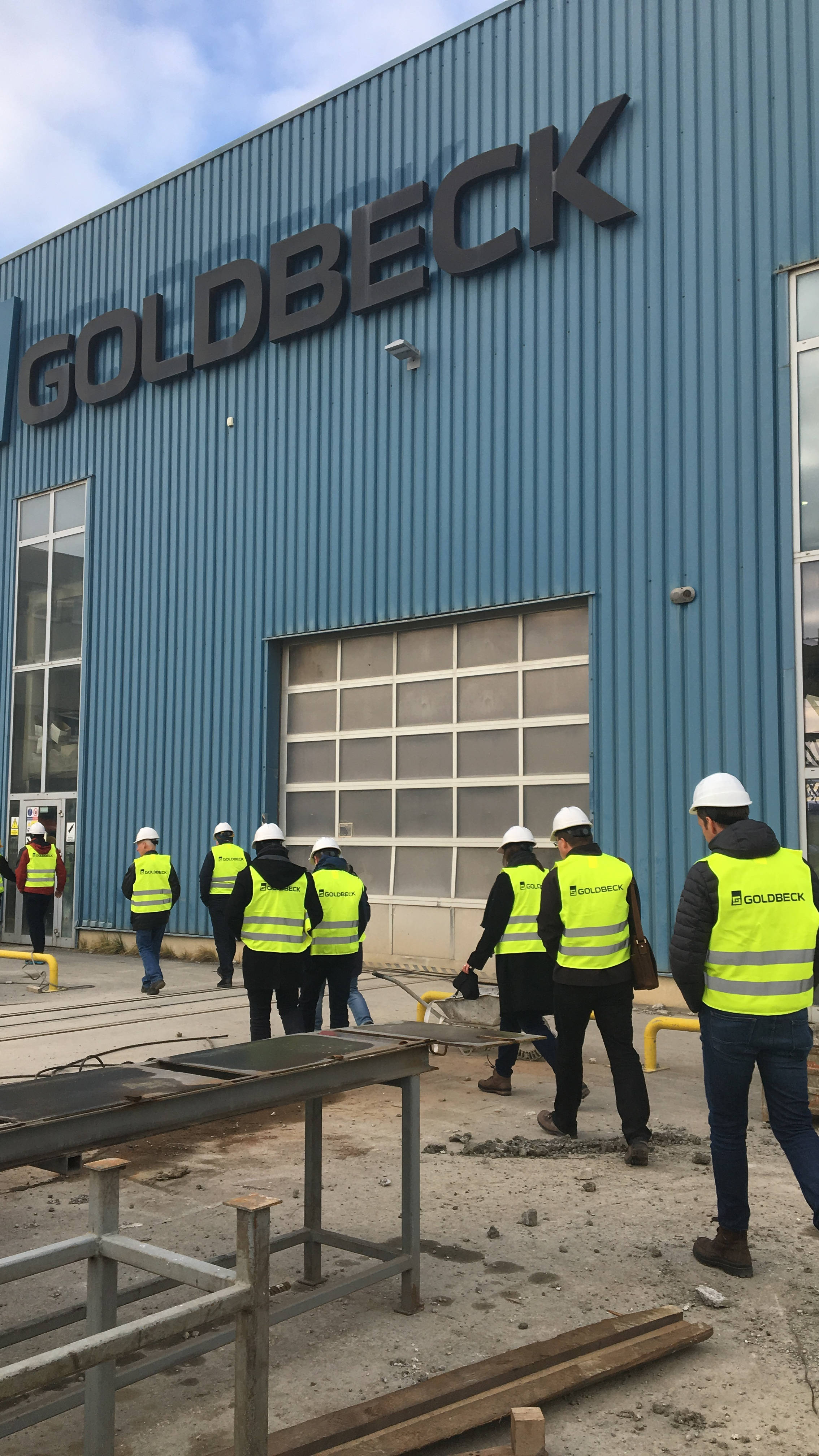 Menschen in Warnwesten und Helmen betreten ein Gebäude mit "GOLDBECK"-Logo, Bauumgebung im Vordergrund.