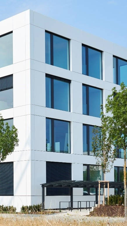 Modernes Bürogebäude mit großer Glasfassade, umgeben von Bäumen und parkenden Autos.