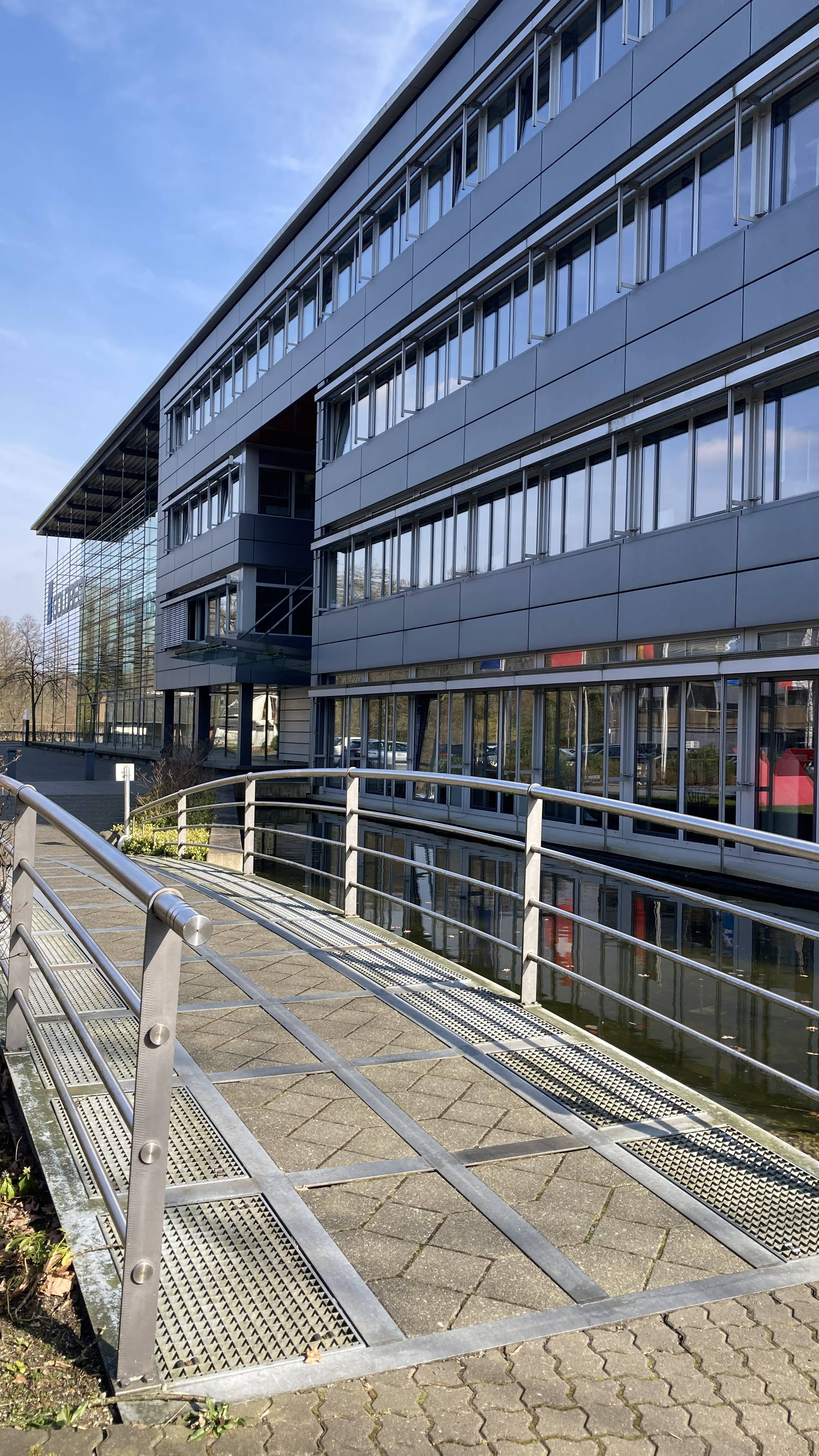 Modernes Bürogebäude mit Glas- und Metallfassade, zugänglich durch eine kleine Brücke über einen Wassergraben.