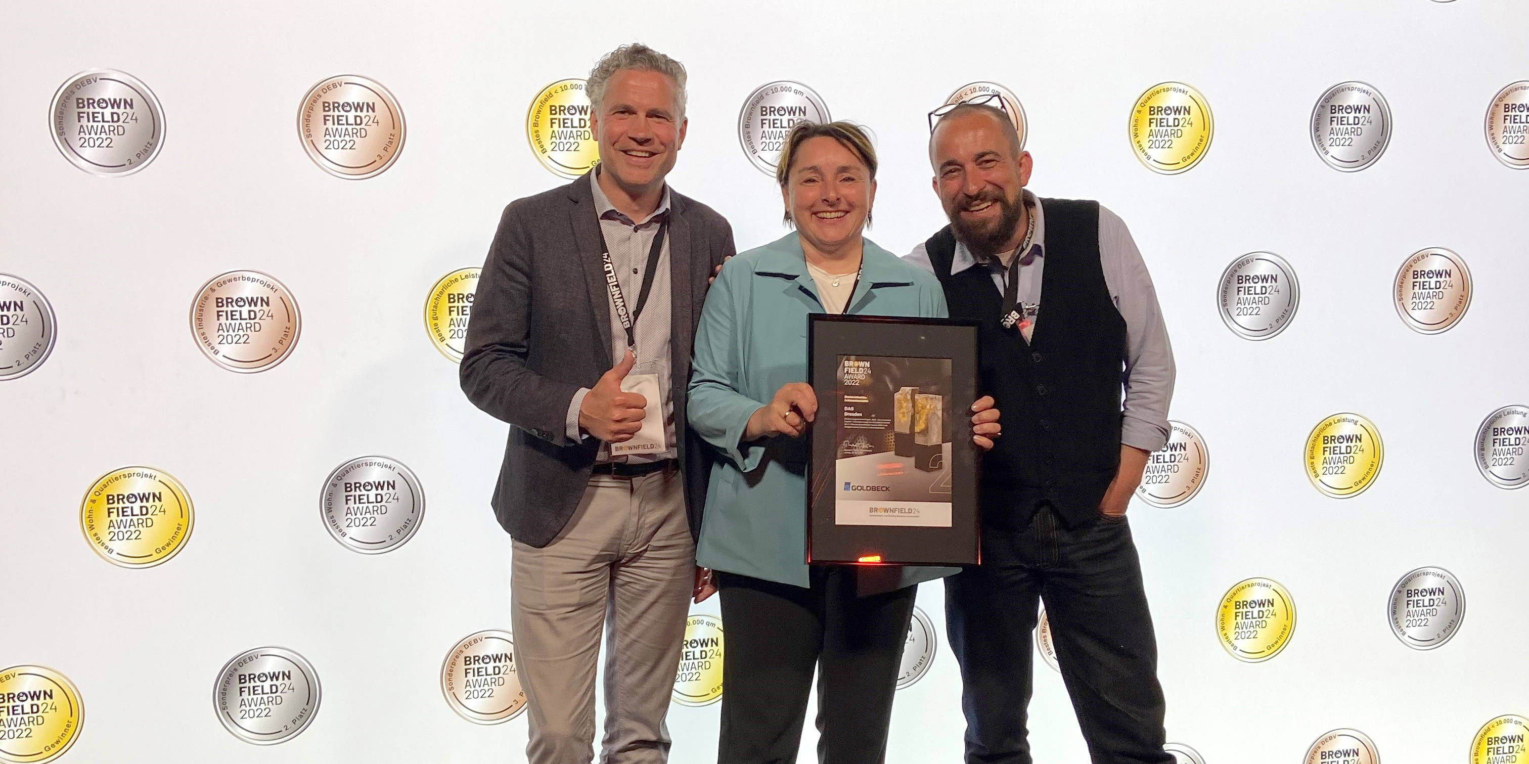 Drei Personen halten eine Auszeichnung vor einer Wand mit "Brown Field24 Award 2022"-Logos und dem Text "Für Schweiß, harte Arbeit und gute Ideen".