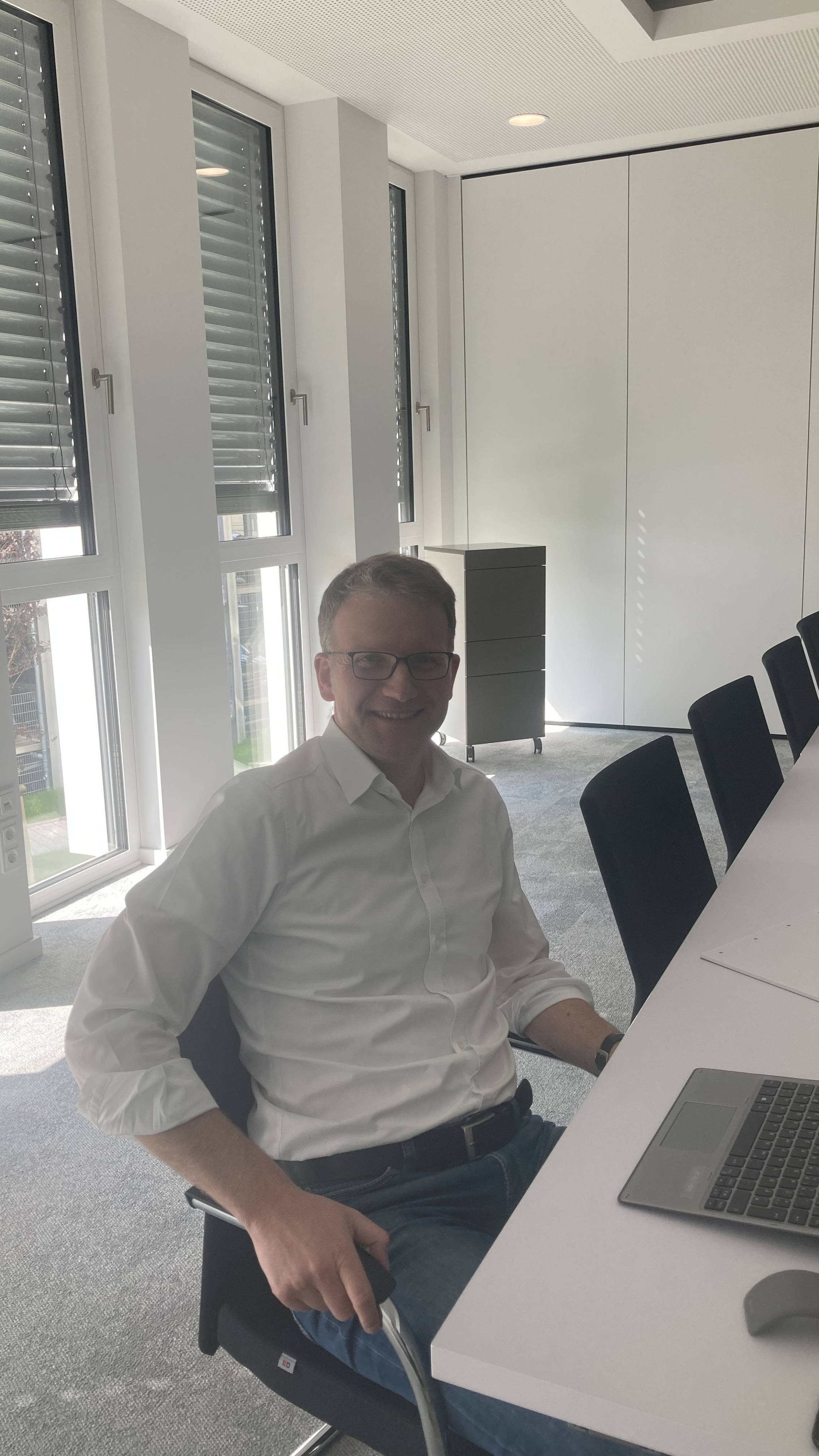 Mann in weißem Hemd sitzt lächelnd an einem Konferenztisch mit Laptop in einem modernen, hellen Büro.