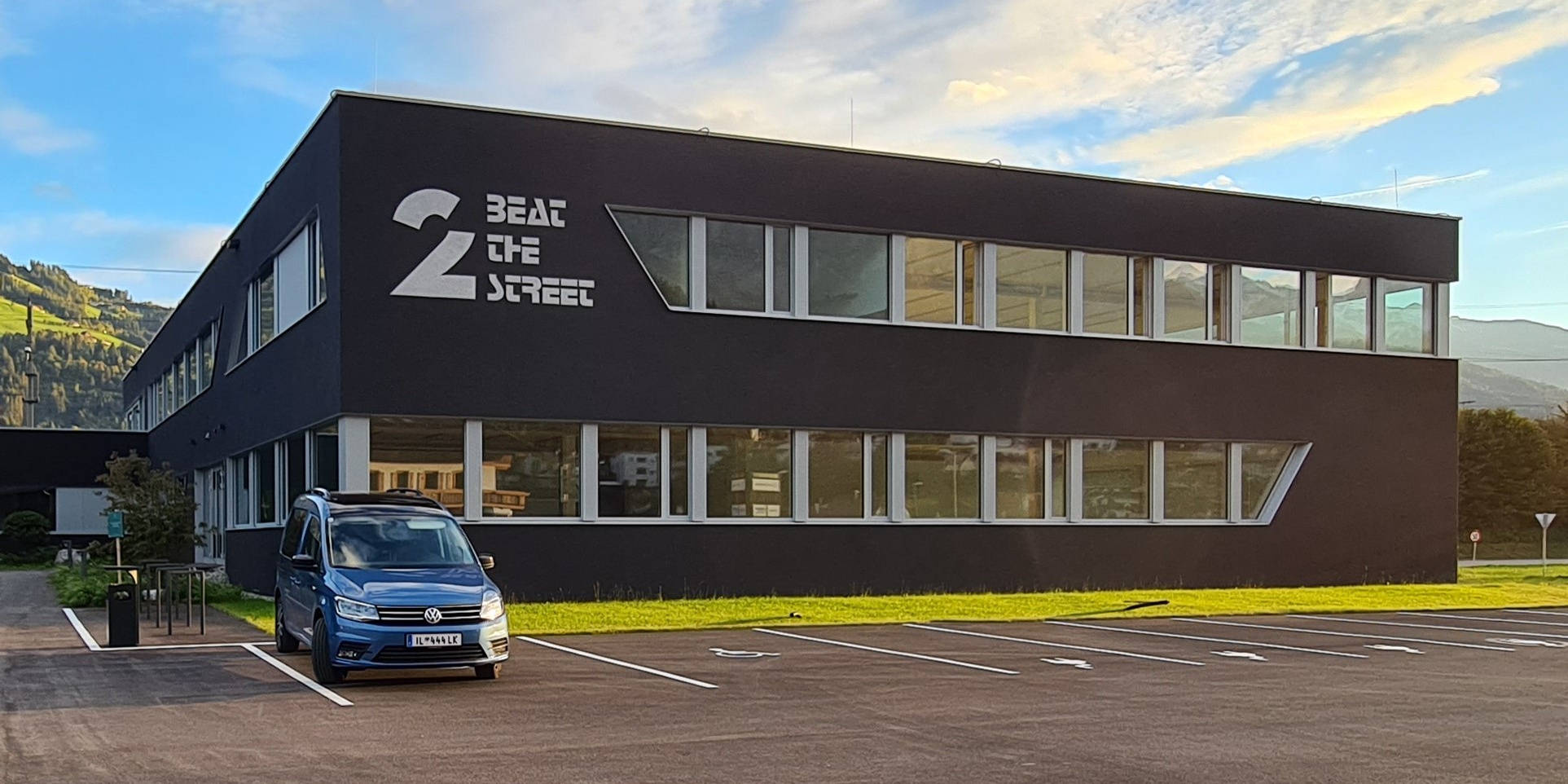 Modernes Gebäude mit der Aufschrift "Beat the Street", daneben ein blauer Van auf einem Parkplatz bei sonnigem Wetter.
