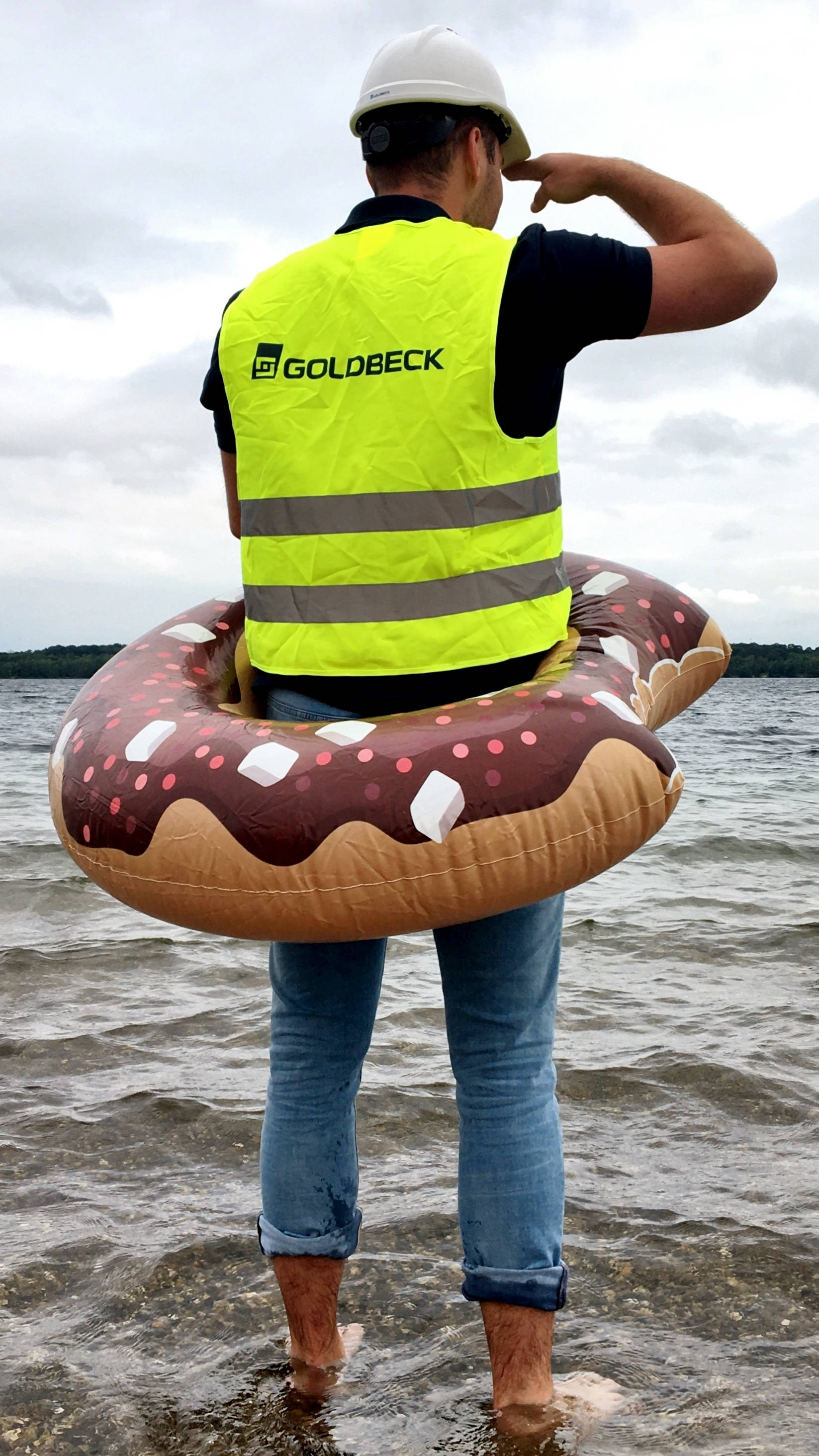 Mann in gelber Sicherheitsweste und Helm mit Donut-Schwimmreifen, steht im flachen Wasser und blickt in die Ferne.