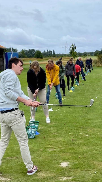 Gruppe von Menschen, die auf einer Driving Range Golf üben, mit Fokus auf den vordersten Spieler in Schwunghaltung.