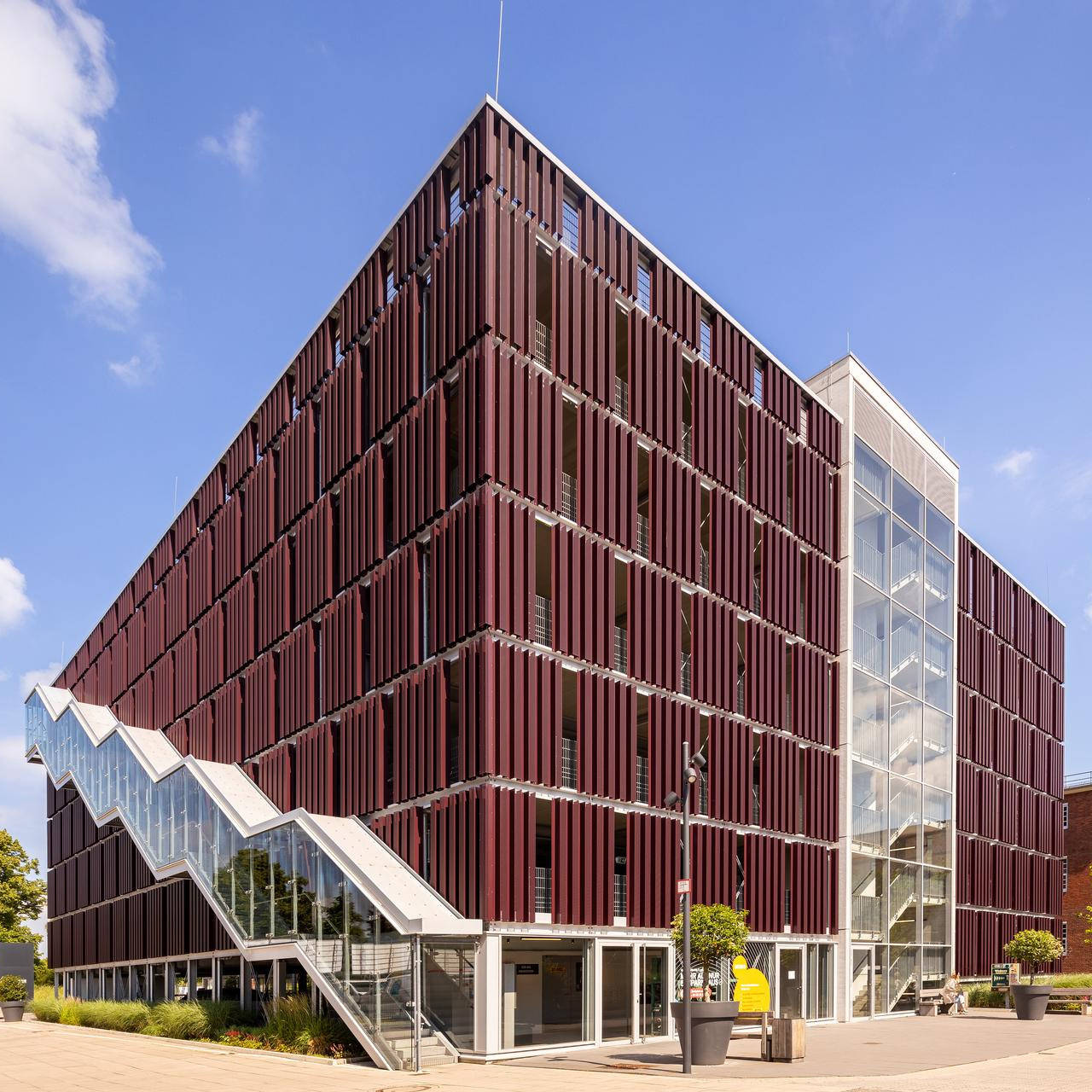 Modernes Bürogebäude mit dunkelroter Fassade, Glasaufzug und Außentreppe, umgeben von Pflanzen bei blauem Himmel.