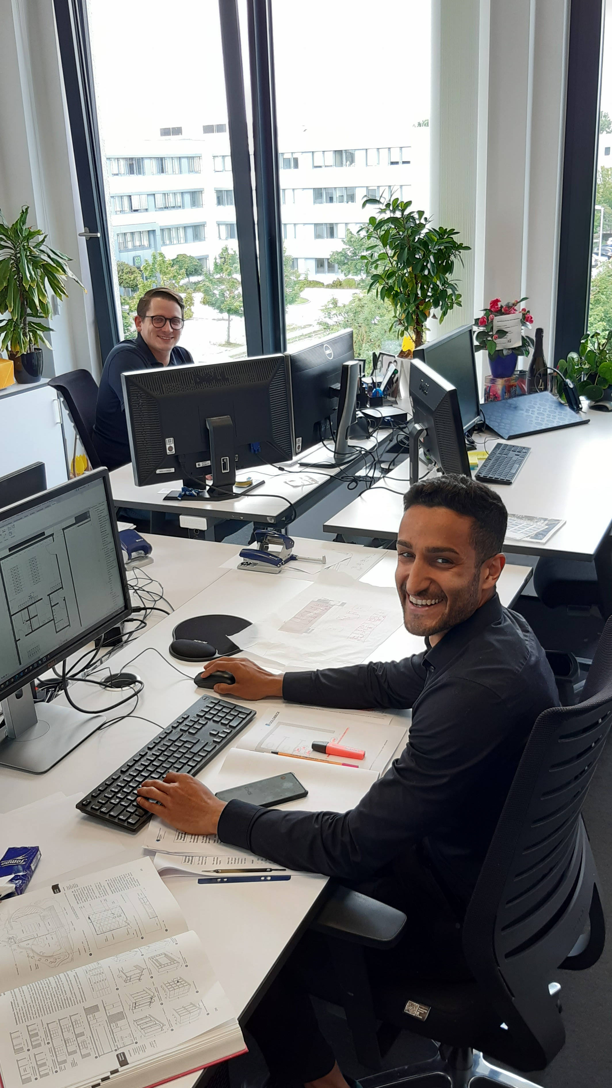 Zwei Männer in einem modernen Büro arbeiten an Computern, umgeben von Pflanzen, mit Stadtblick durch große Fenster.