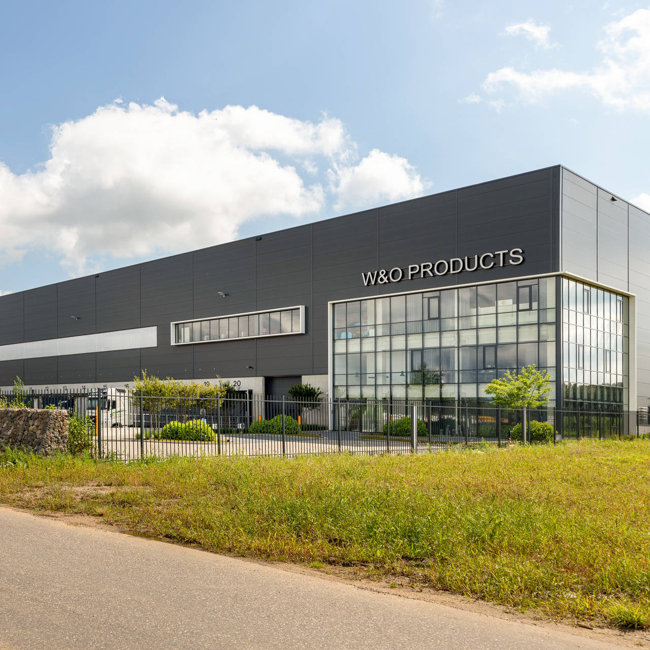 Exterieur van een groot, modern bedrijfsgebouw met de naam "W&O Products" op de gevel. Er is een goed onderhouden grasveld en een omheining aan de voorkant. De lucht is helder met enkele wolken. Op de voorgrond staat een bord met bedrijfsnamen.