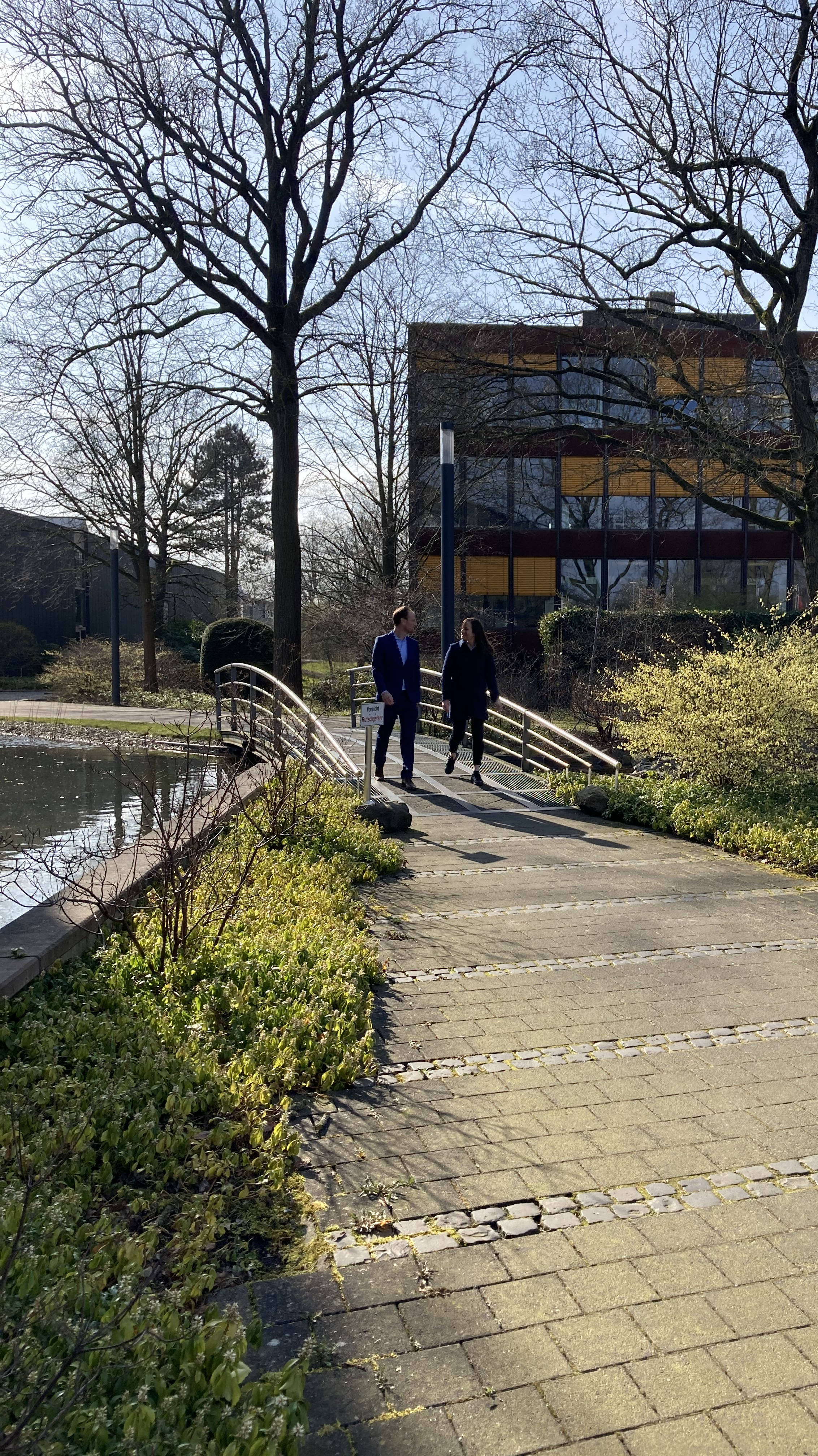 Zwei Personen gehen auf einem gepflasterten Weg über eine kleine Brücke in einem Park, im Hintergrund ein modernes Gebäude und Bäume.