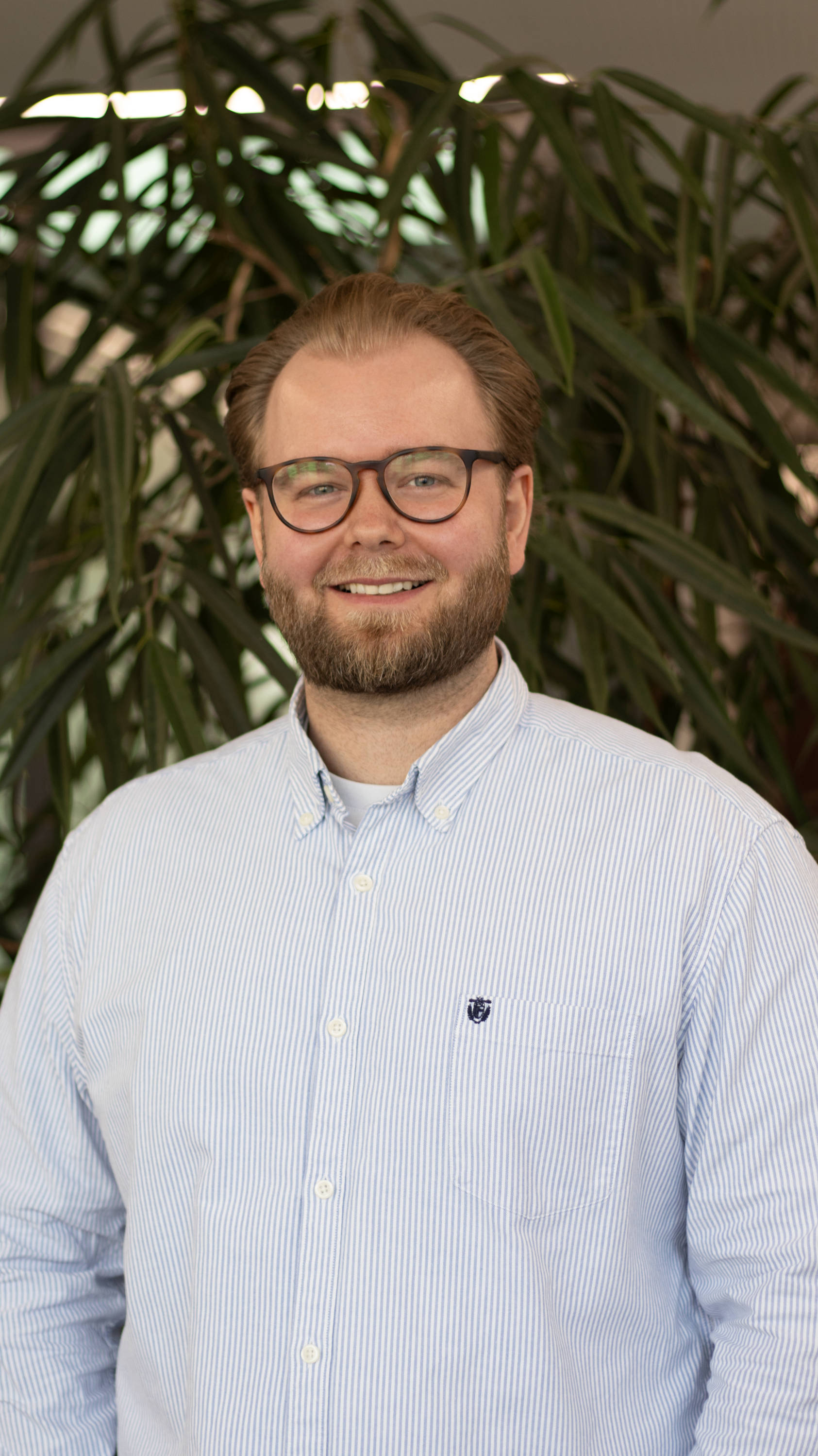 Mann mit Brille und Bart lächelt vor Pflanzenhintergrund. Trägt ein gestreiftes Hemd.