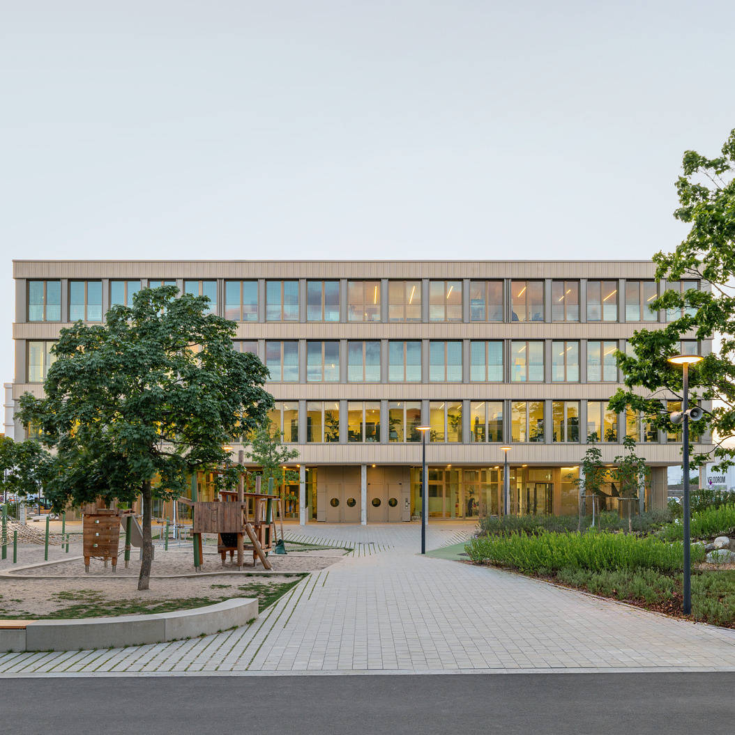 Moderne Schule mit vier Etagen, umgeben von Bäumen und einem Spielplatz im Vordergrund.