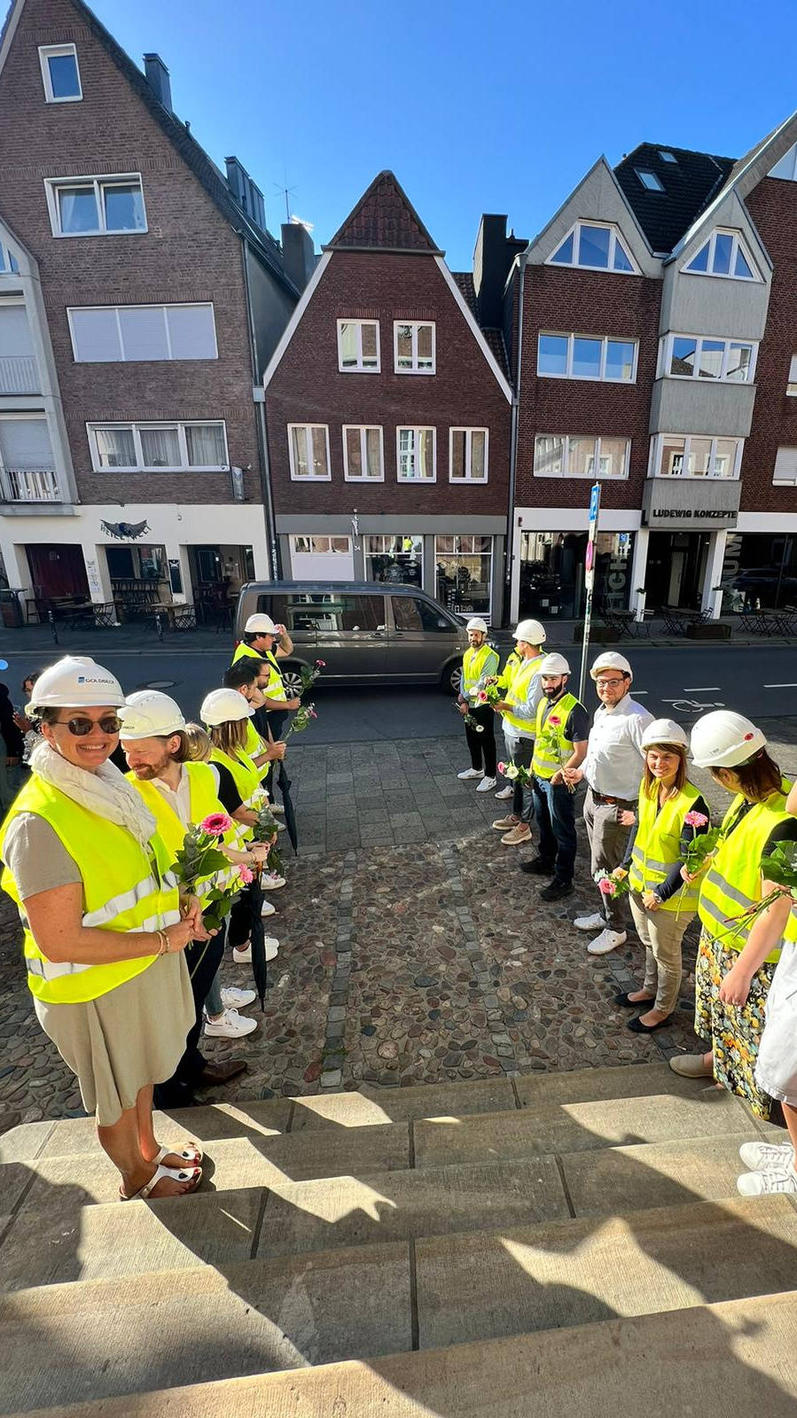 Gruppe von Menschen in gelben Warnwesten und Helmen steht mit Blumen in den Händen auf einer Treppe vor historischen Häusern.