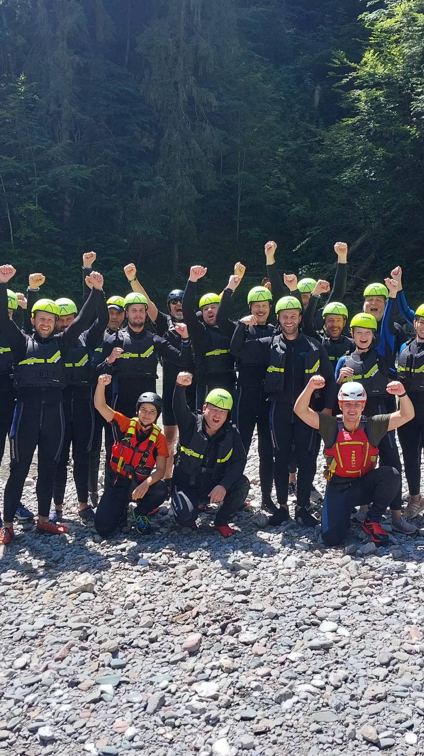 Gruppe von Menschen in Neoprenanzügen und Helmen posiert jubelnd am Flussufer während eines Outdoor-Abenteuers.