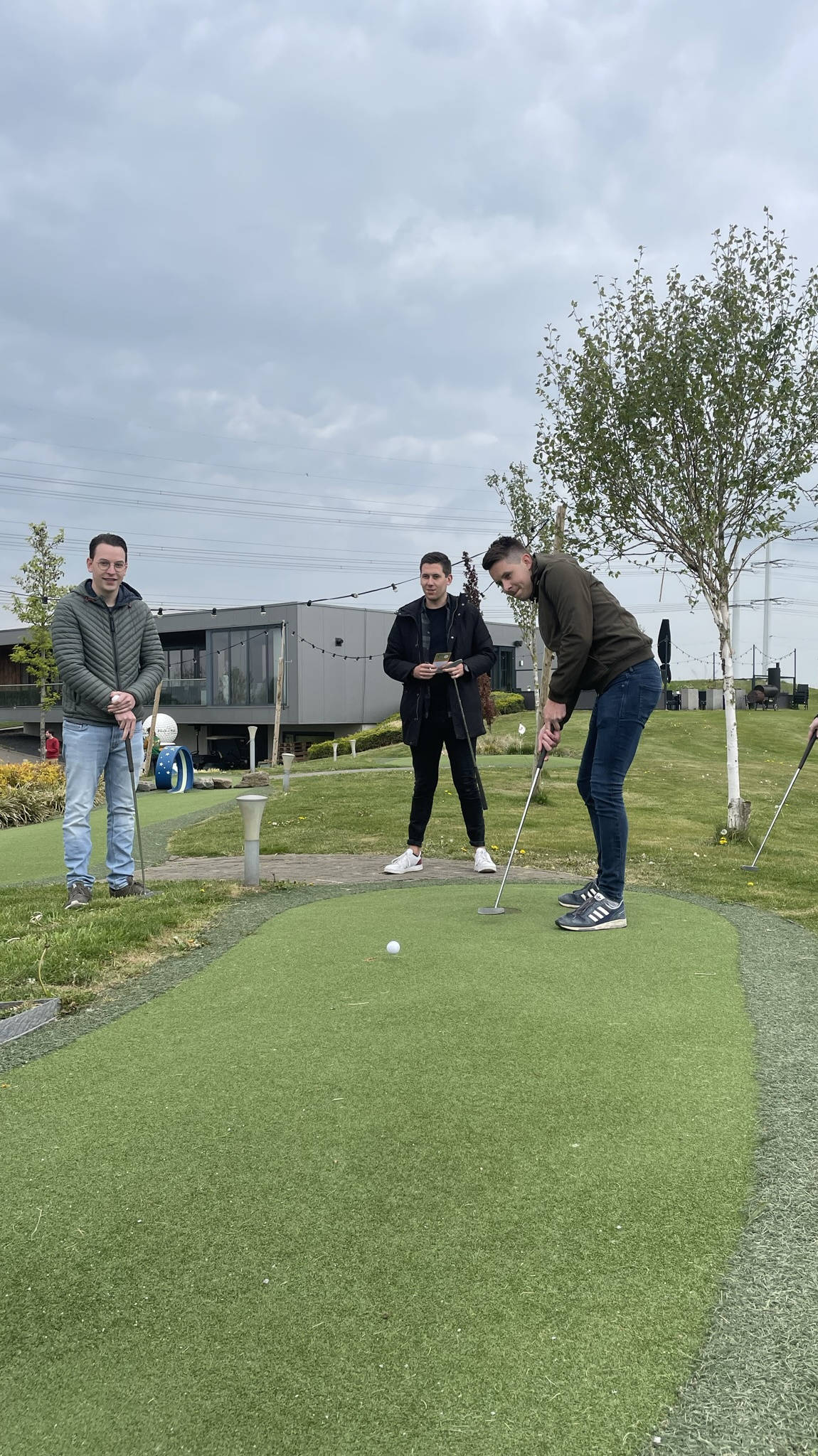 Vier Männer spielen Minigolf auf einem Außenplatz unter bewölktem Himmel.