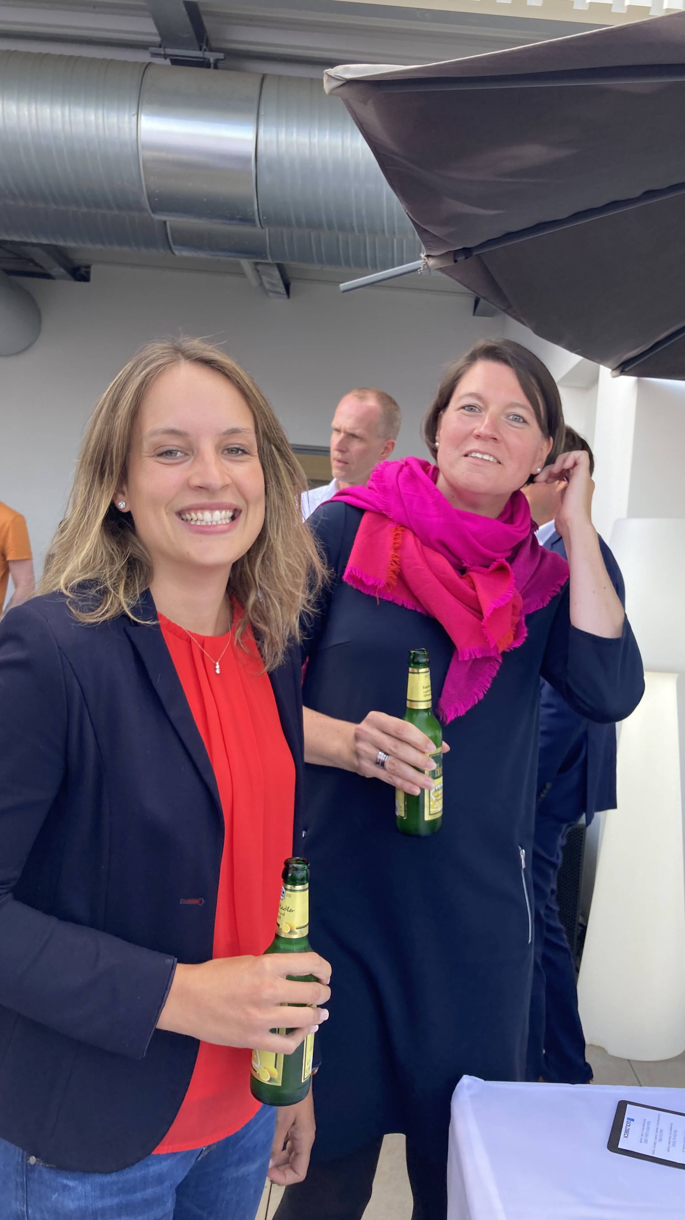 Zwei lächelnde Frauen bei einem Event im Freien halten Bierflaschen in der Hand, unter einem großen Sonnenschirm.