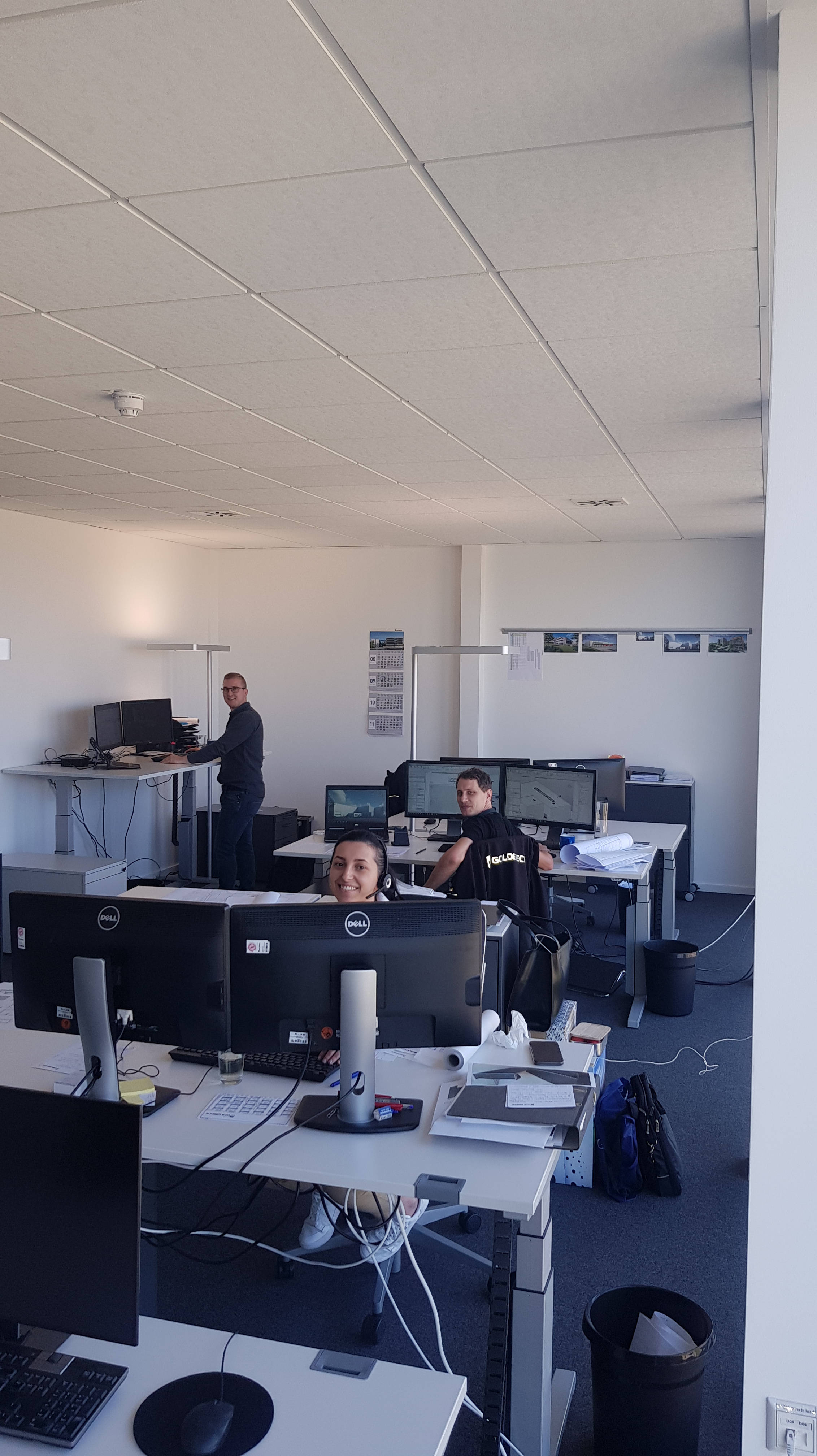 Büro mit mehreren Mitarbeitern an Computern, zwei Personen sitzen, eine steht an einem höhenverstellbaren Schreibtisch.