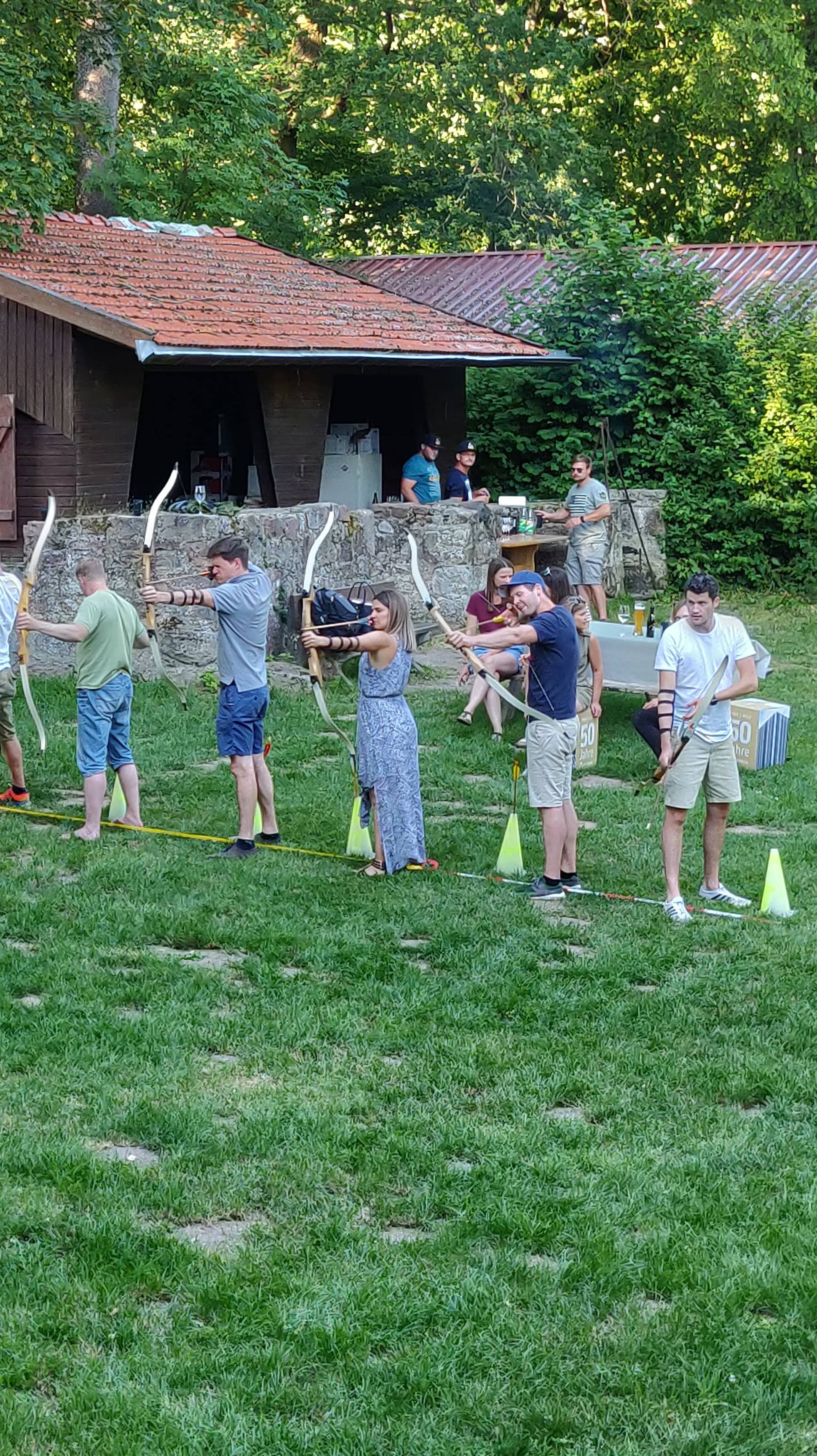 Gruppe von Menschen übt Bogenschießen auf einer Wiese vor einer Holzhütte.