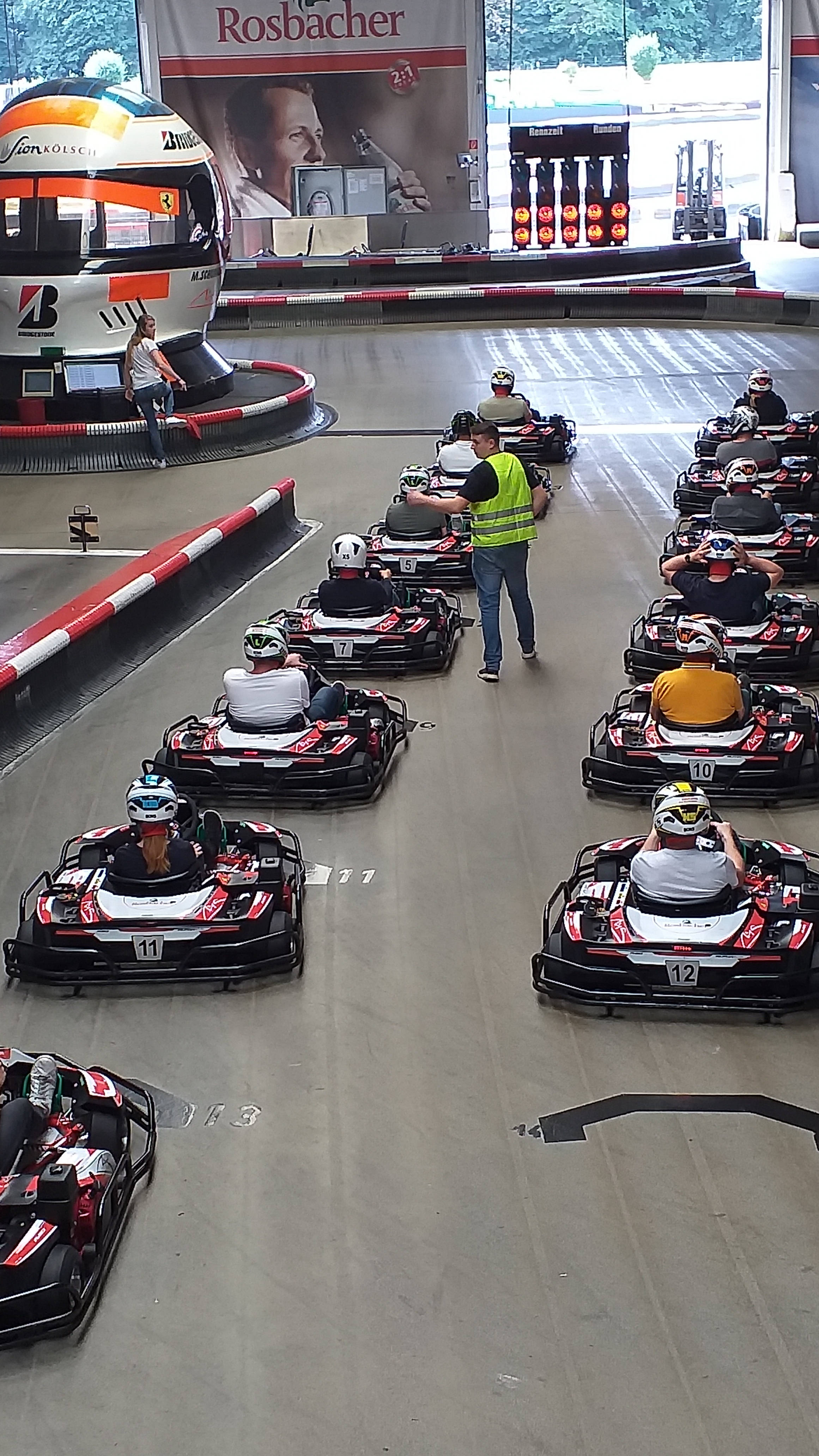 Kartfahrer in einer Indoor-Kartbahn, bereit für ein Rennen. Ein Streckenwart in gelber Weste gibt Anweisungen.