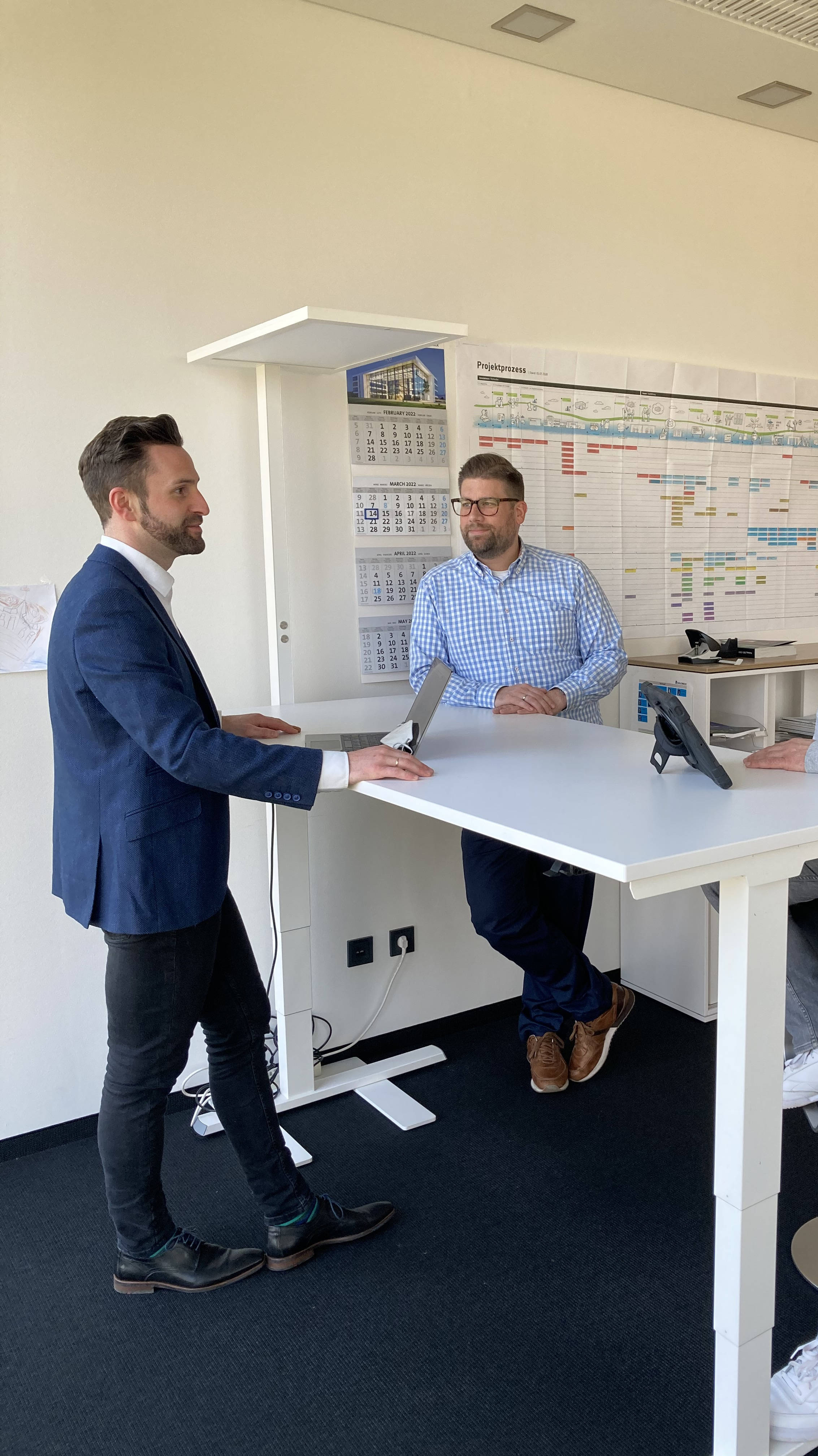 Drei Männer bei einem Meeting an einem Stehtisch im Büro, mit Projektplan an der Wand im Hintergrund.
