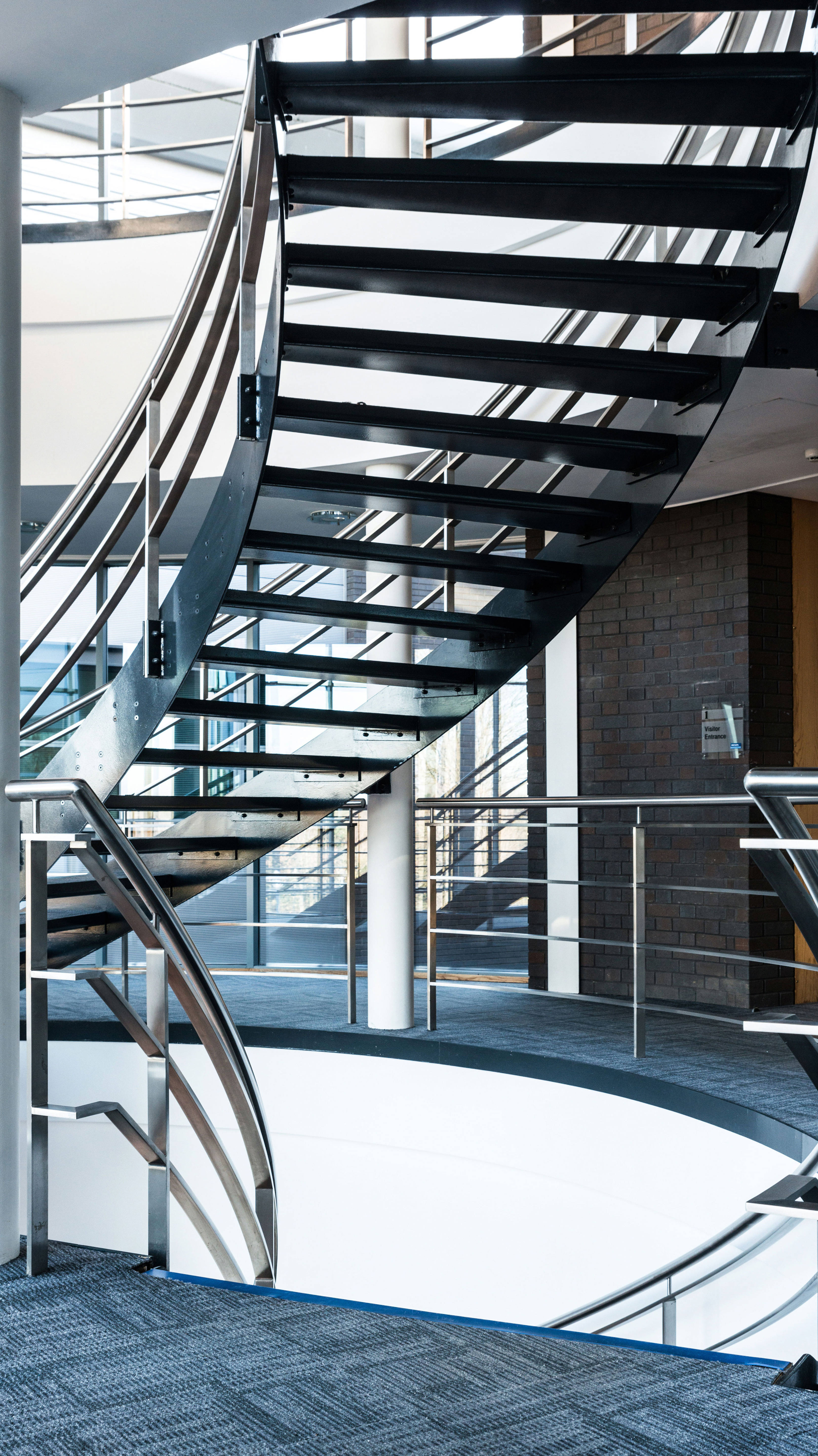 Moderne Wendeltreppe aus Metall in einem Bürogebäude mit Teppichboden und Glasgeländern.
