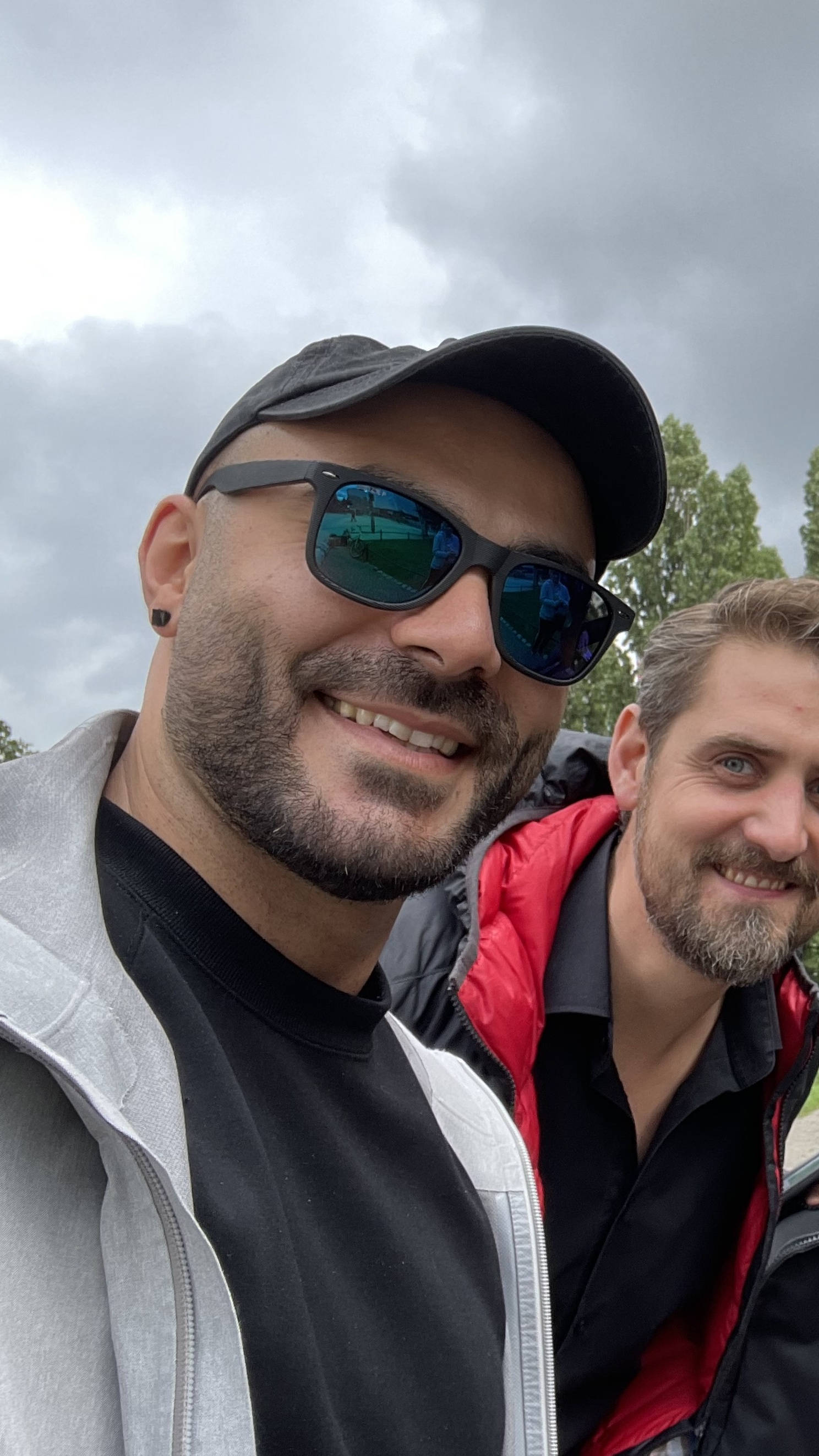 Two men smiling at the camera, one wearing sunglasses and a baseball cap. Background with trees and cloudy sky.