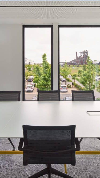 Konferenzraum mit leerem Tisch und Stühlen, großem Fensterblick auf einen Industriekomplex und grüner Umgebung.