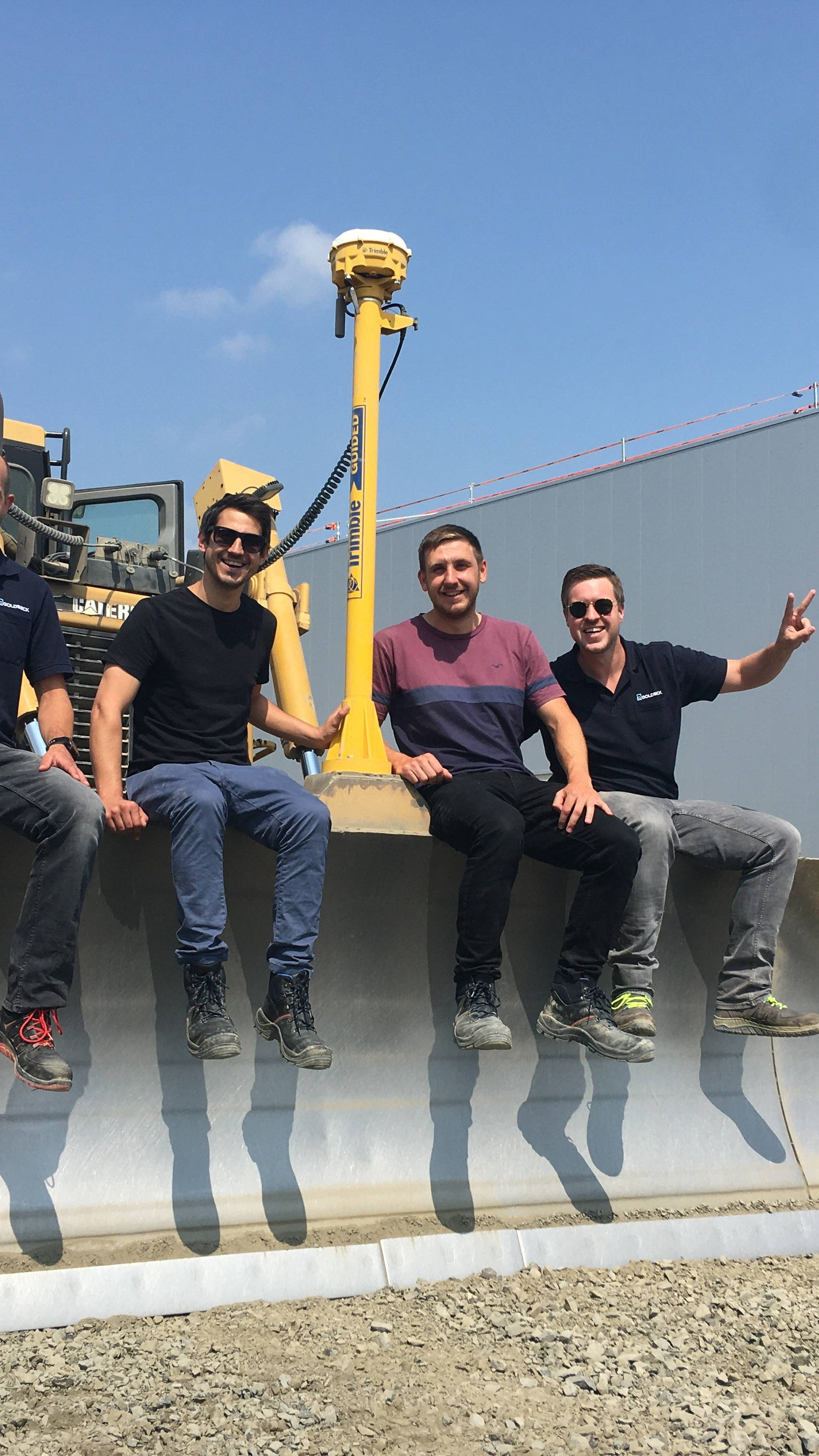 Vier Männer sitzen auf der Schaufel eines gelben Bulldozers auf einer Baustelle, lächeln und zeigen Handzeichen.