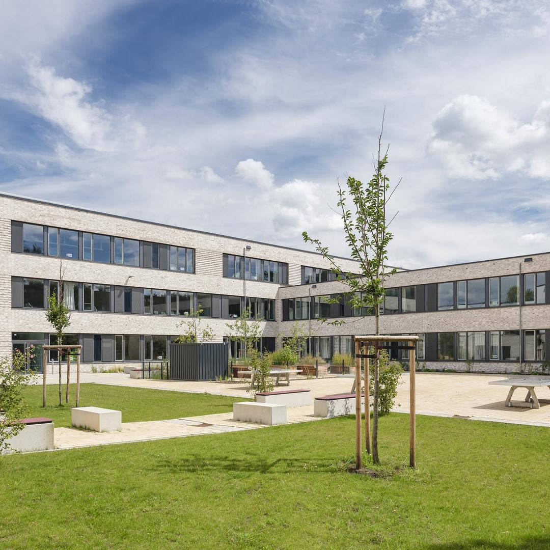 Modernes Schulgebäude mit großer Fensterfront, umgeben von grünem Schulhof und Gehwegen bei sonnigem Wetter.