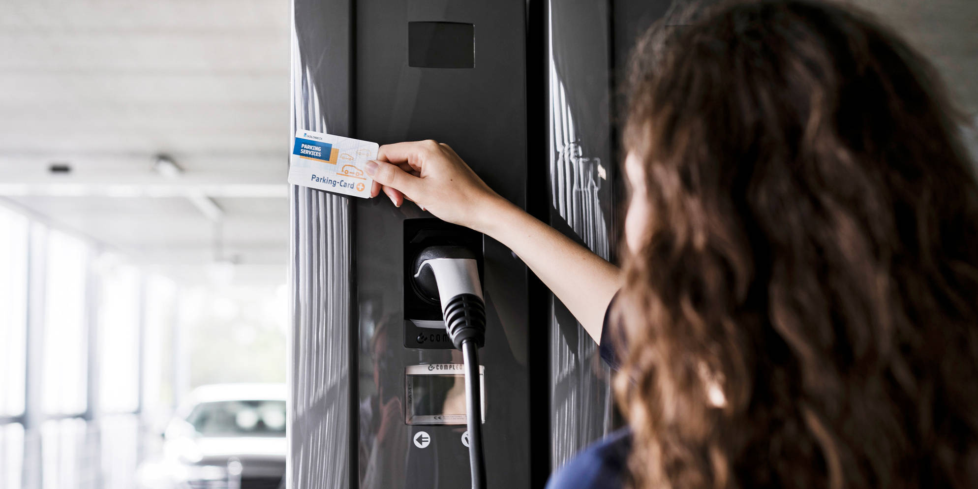 Eine Frau mit braunen Haaren hält die Goldbeck Parking Card an eine Ladesäule.