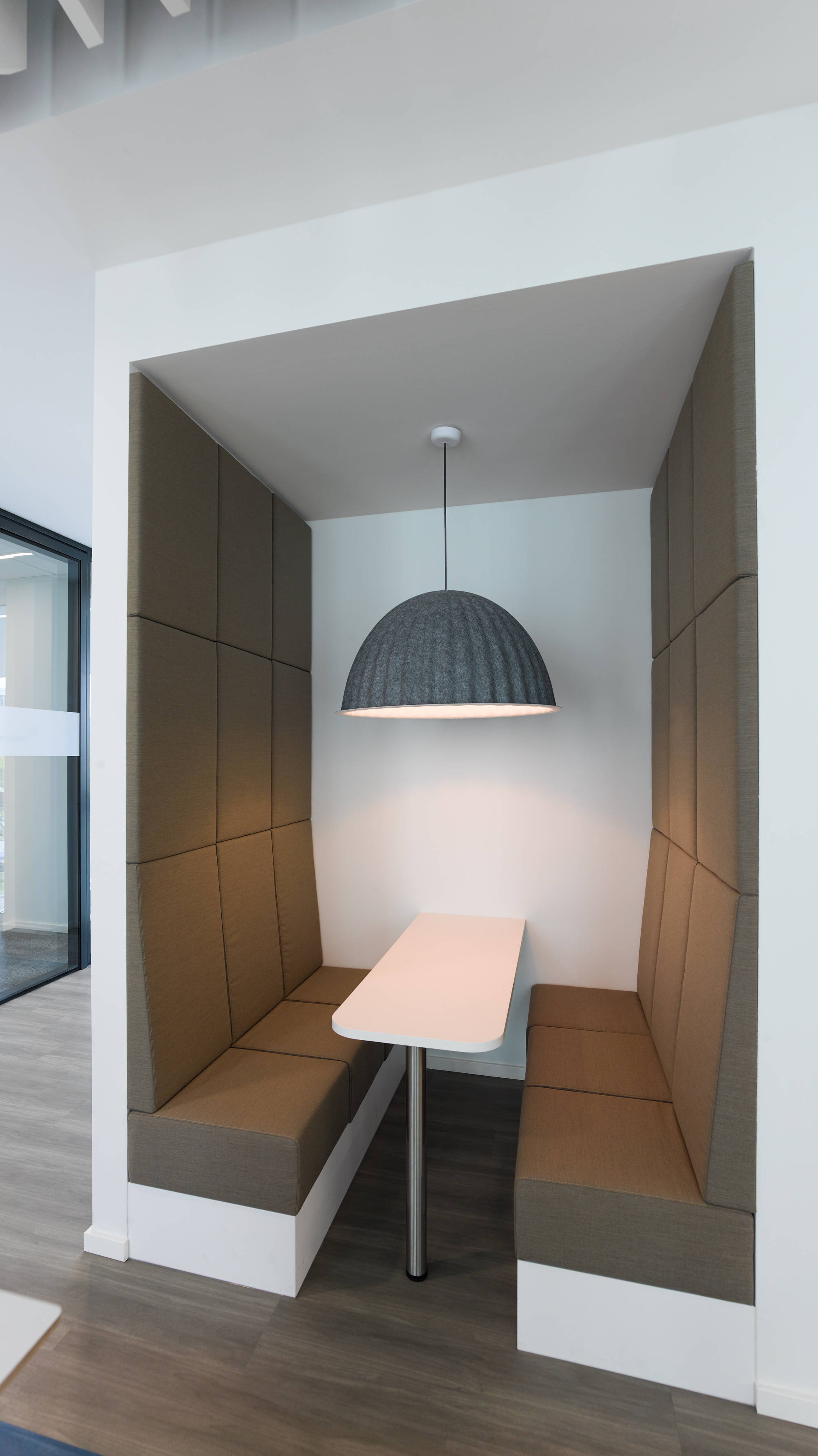 Moderne Büro-Nische mit gepolsterten Sitzen, weißem Tisch und hängender Lampe, ideal für Meetings.