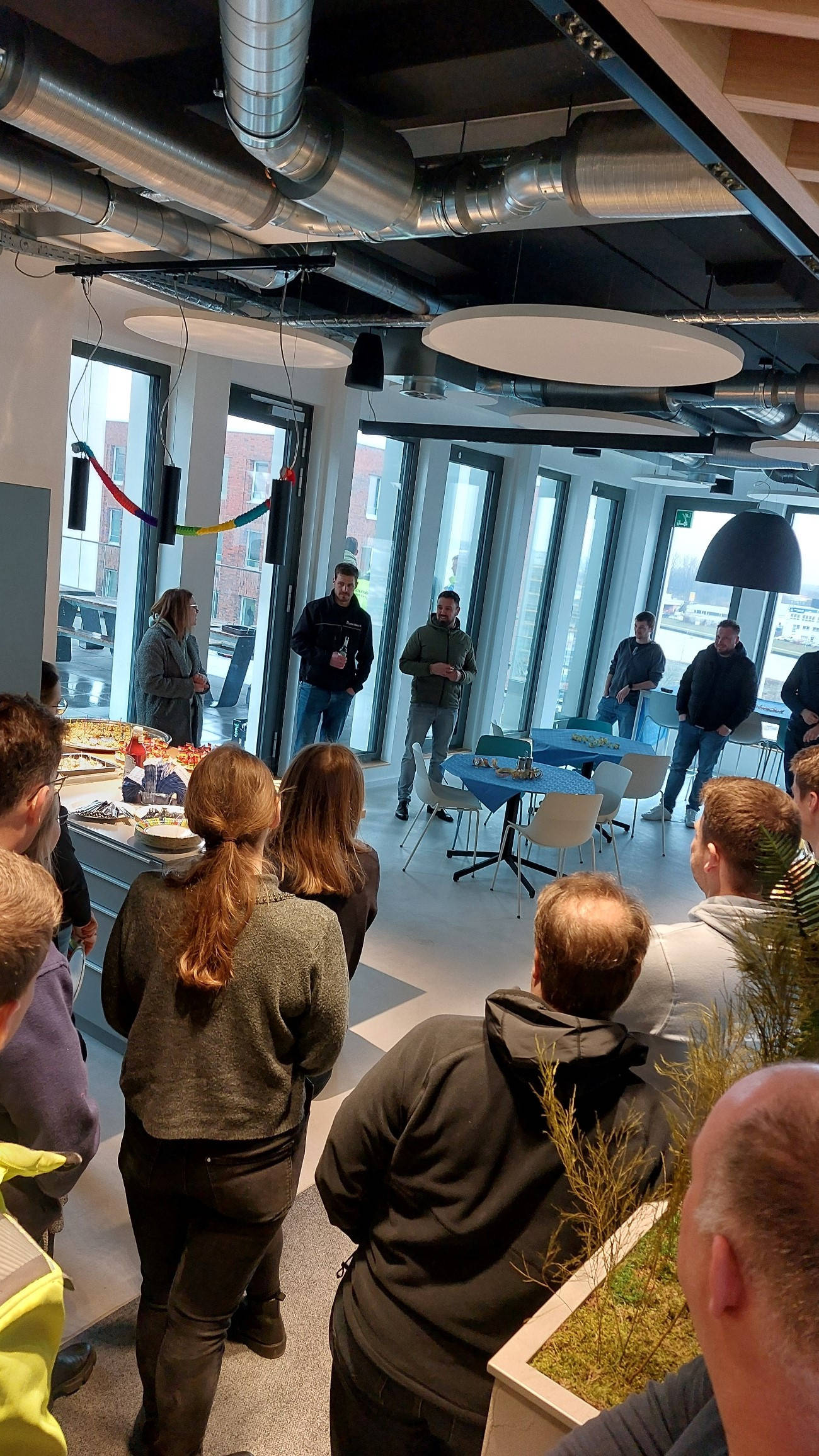 Versammlung von Mitarbeitern in einem modernen Büro, die einer Präsentation zuhören. Im Hintergrund sind große Fenster und Dekorationen zu sehen.