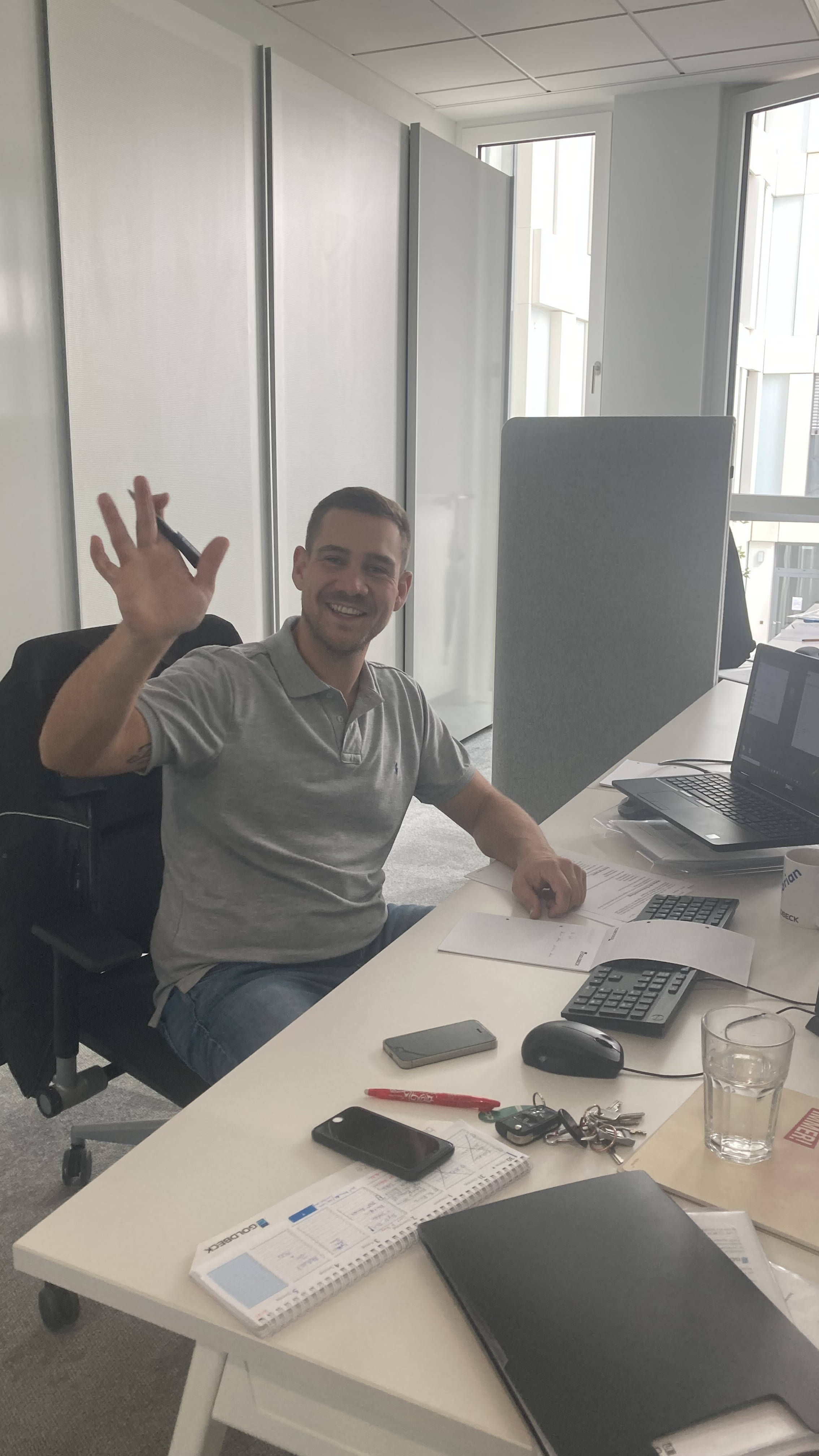 Mann in grauem Polohemd lächelt und winkt am Schreibtisch im modernem Büro, vor Laptop und Dokumenten.