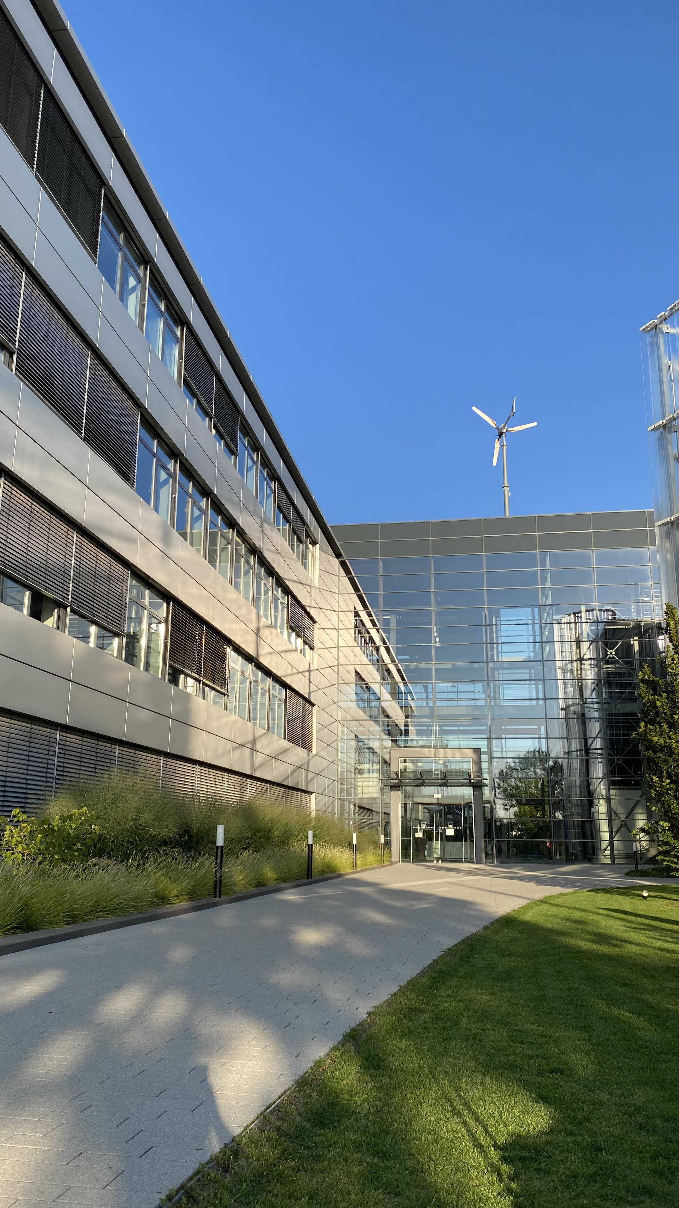 Moderner Bürogebäudekomplex mit Glasfassade und Windturbine auf dem Dach, umgeben von Gartenanlagen und beleuchtetem Weg.