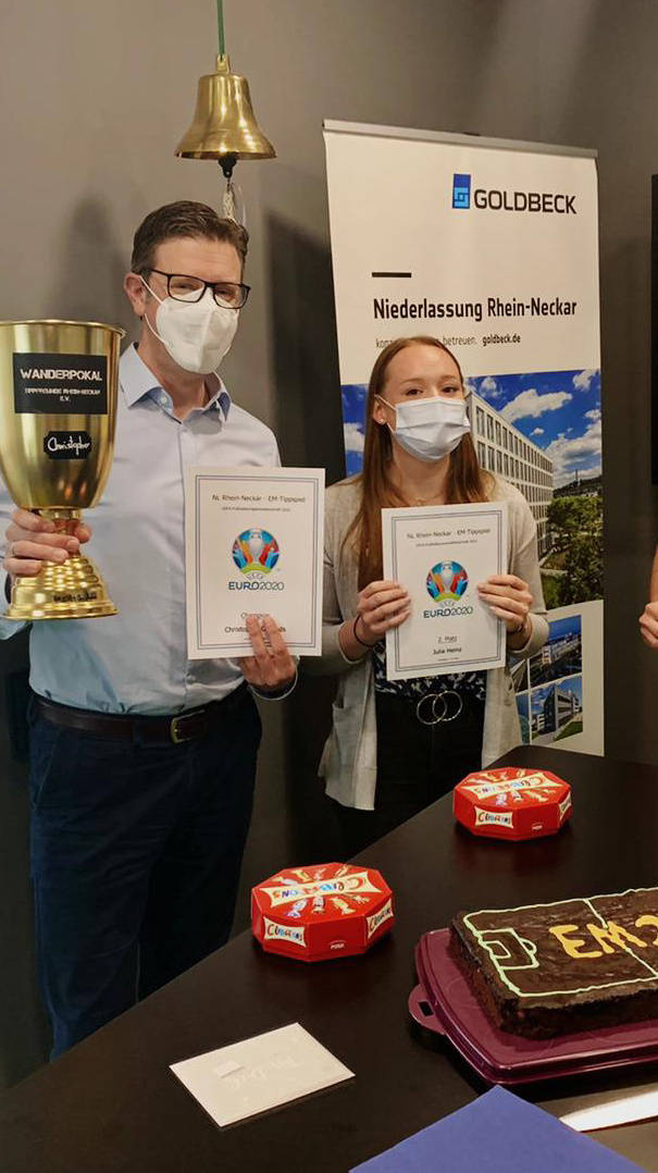 Drei Personen mit Gesichtsmasken halten Urkunden und einen Pokal bei einer Preisverleihung in einem Büro von Goldbeck. Vor ihnen ein Kuchen mit der Aufschrift "EM 2020".