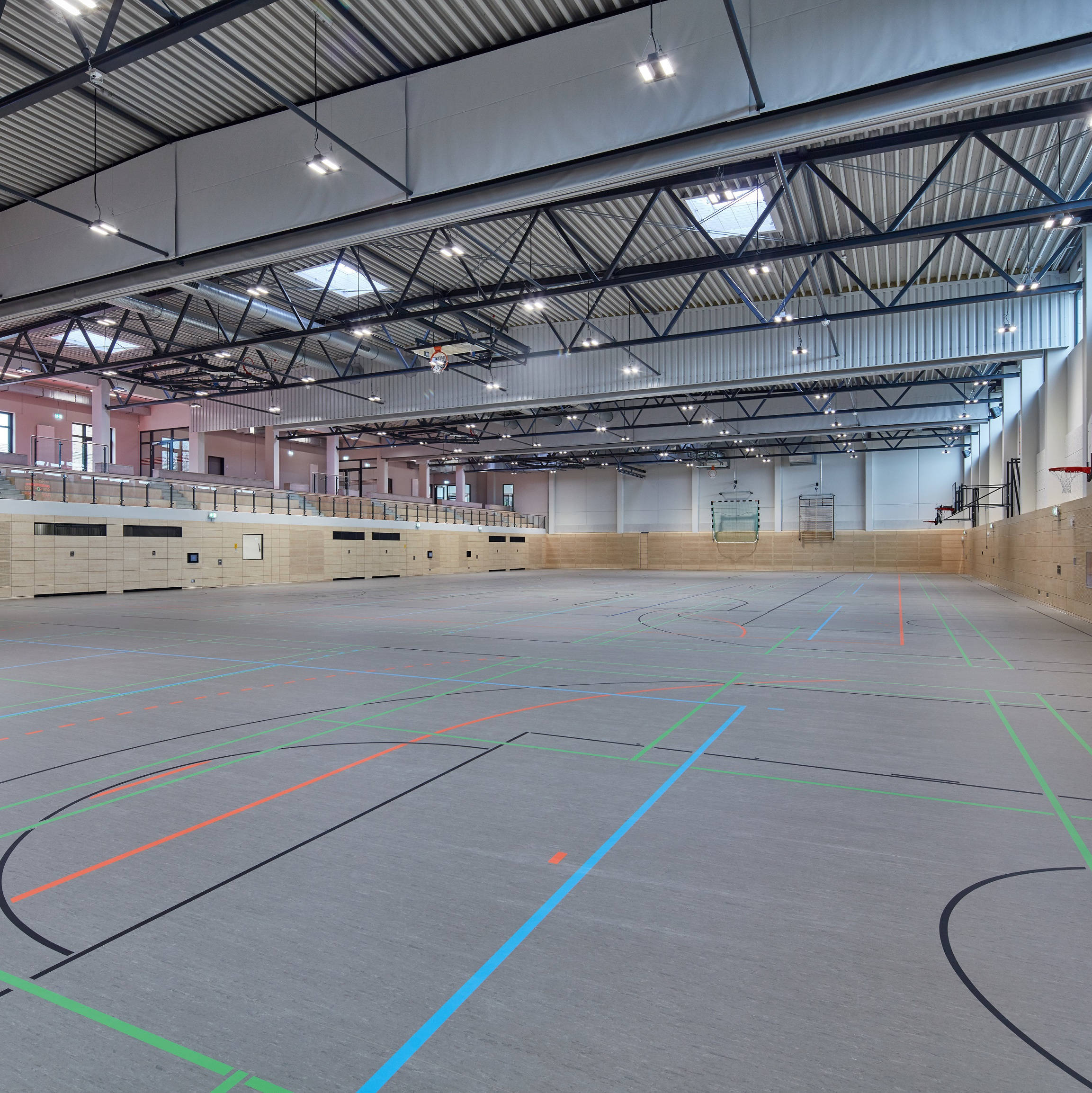Große, leere Sporthalle mit markierten Spielfeldern auf dem Boden und hohen Deckenstrukturen.