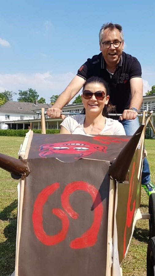 Man and woman drive across a meadow on a sunny day in a self-built soapbox car.