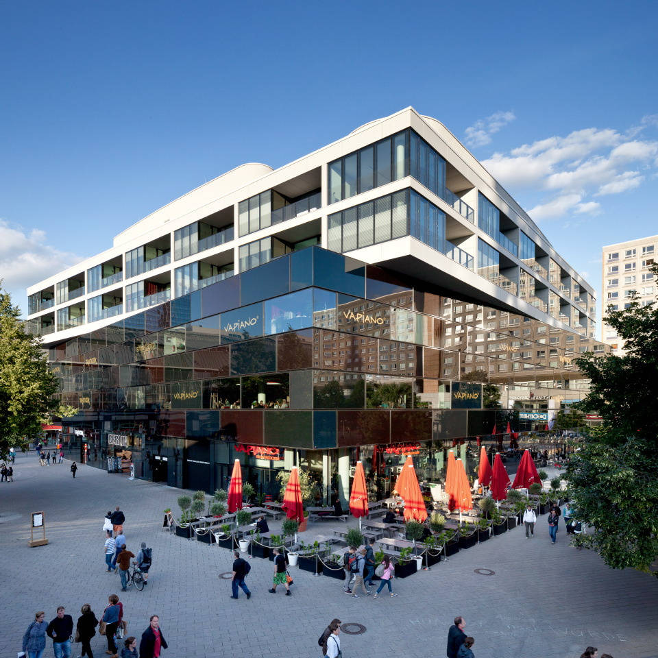 Modernes Gebäude mit Glasfassade und Restaurantterrasse, umgeben von Fußgängern und Bäumen.
