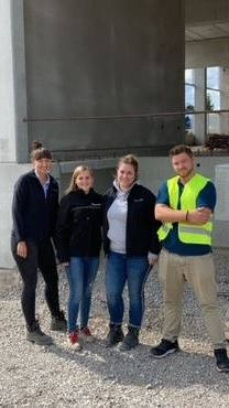 Gruppe von vier Personen in Freizeitkleidung und Warnweste auf einer Baustelle vor einem Betongebäude.