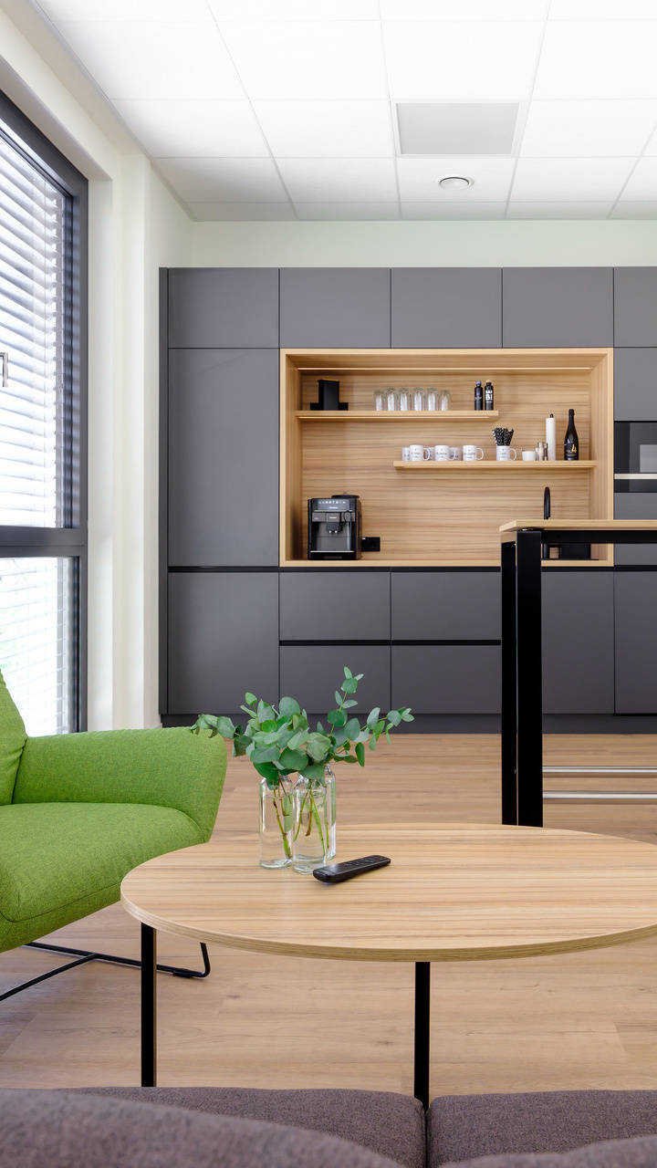 Moderne Büro-Küche mit grauen Schränken, grünem Lounge-Sessel, kleinem Rundtisch mit Pflanze und Laptop auf Stehtisch.