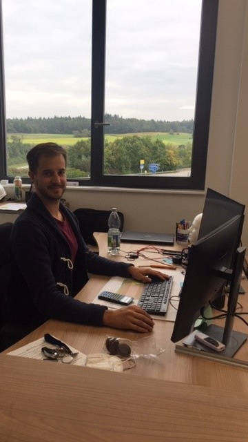 Ein Mann sitzt lächelnd an einem Schreibtisch im Büro und arbeitet am Computer, während er aus dem Fenster ins Grüne blickt.