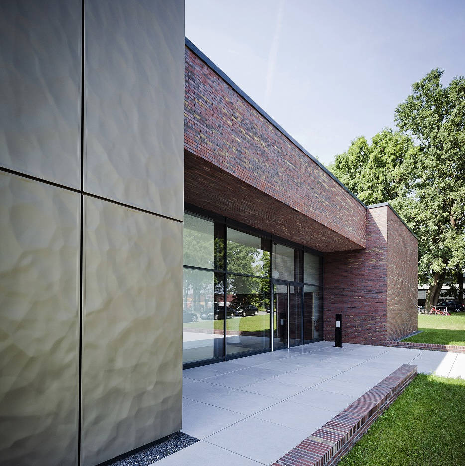 Moderner Eingangsbereich eines Gebäudes mit Glasfront und Klinkerfassade, daneben gepflegte Grünfläche.