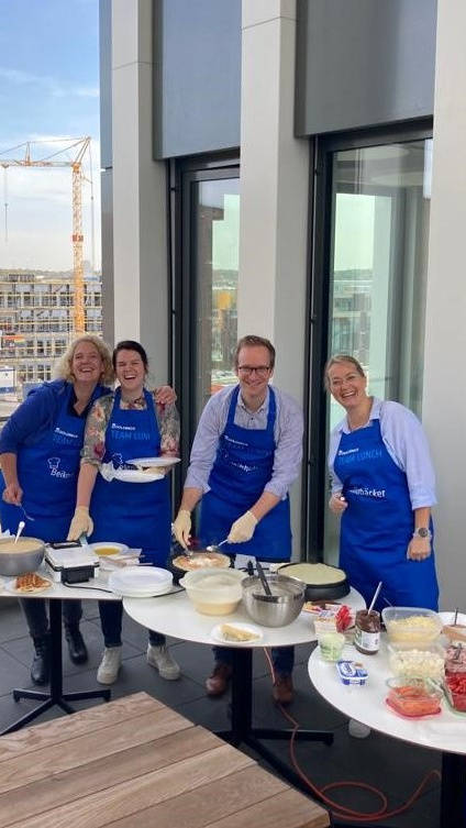 Vier Personen in blauen Schürzen lachen gemeinsam, während sie auf einer modernen Terrasse Kochaktivitäten ausführen. Im Hintergrund ist eine Baustelle zu sehen.