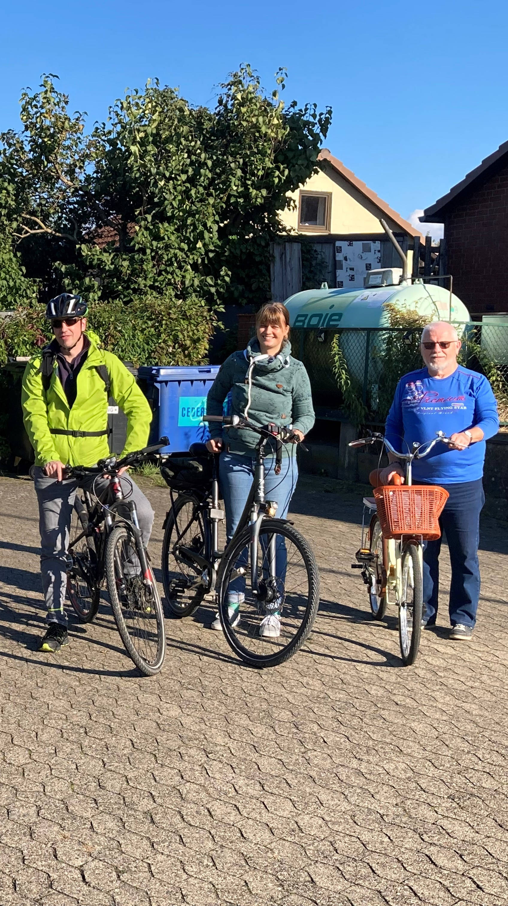 Gruppe von sechs Personen mit Fahrrädern, stehend auf einem gepflasterten Platz bei sonnigem Wetter, umgeben von Bäumen und Häusern.