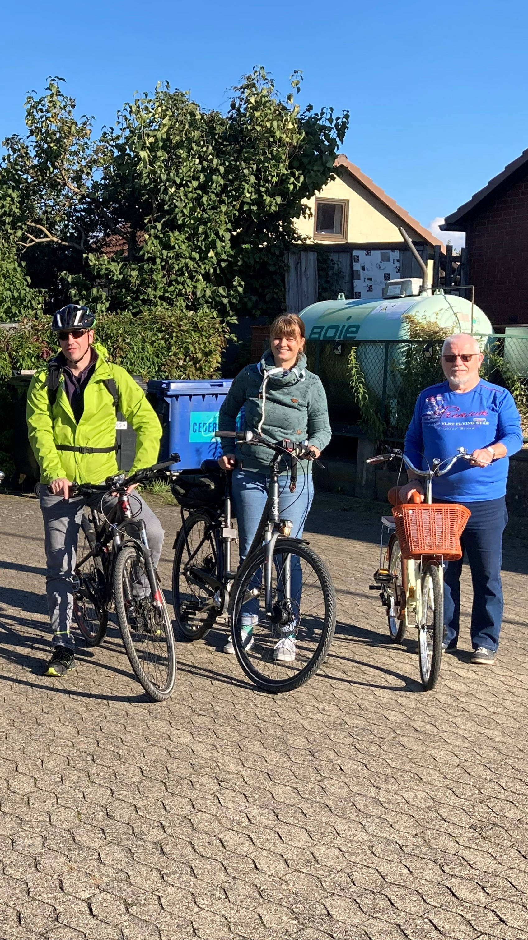 Gruppe von sechs Personen mit Fahrrädern, stehend auf einem gepflasterten Platz bei sonnigem Wetter, umgeben von Bäumen und Häusern.