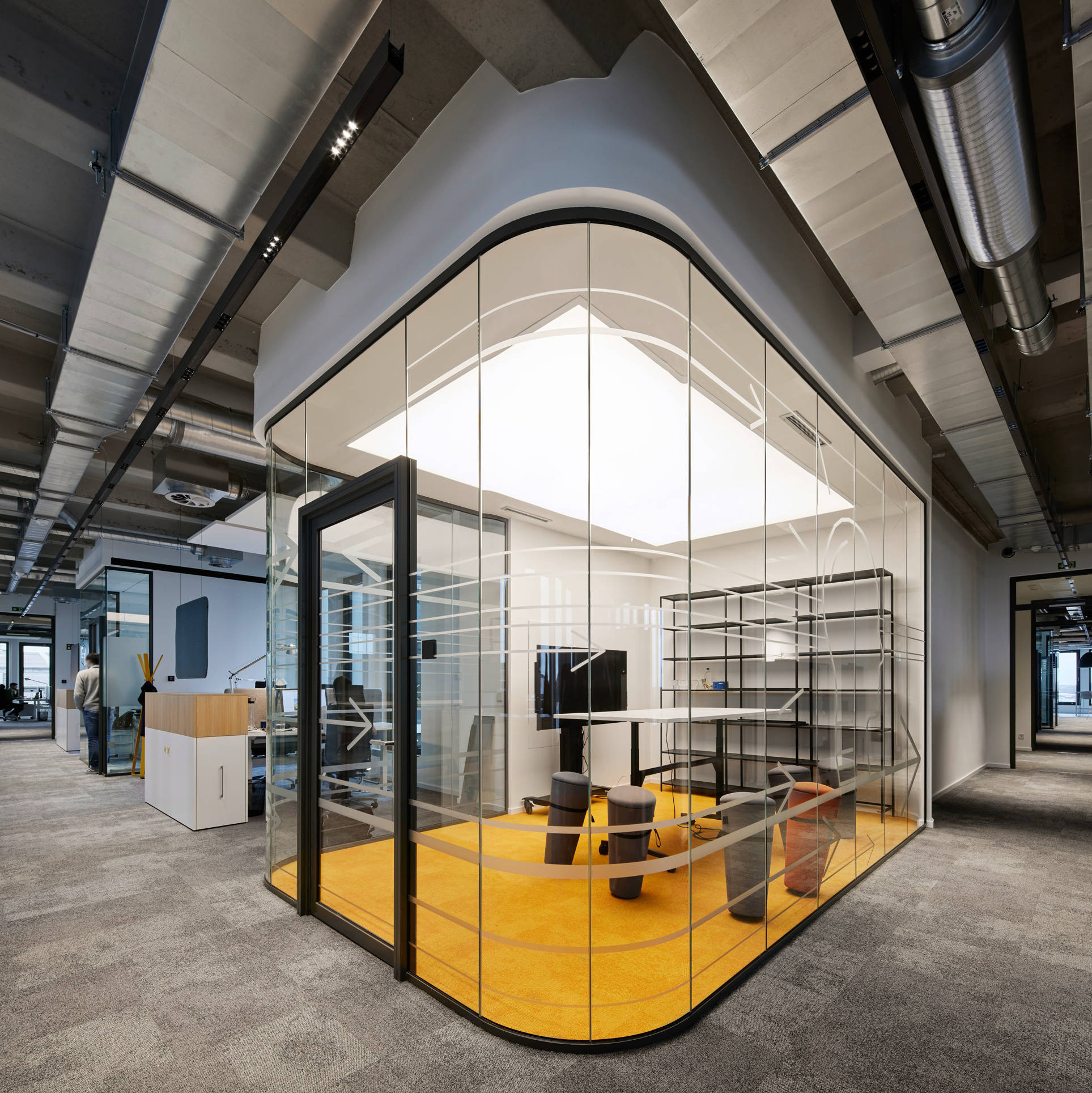 Modern office space with glass partition wall and yellow carpet, surrounded by desks and shelves.