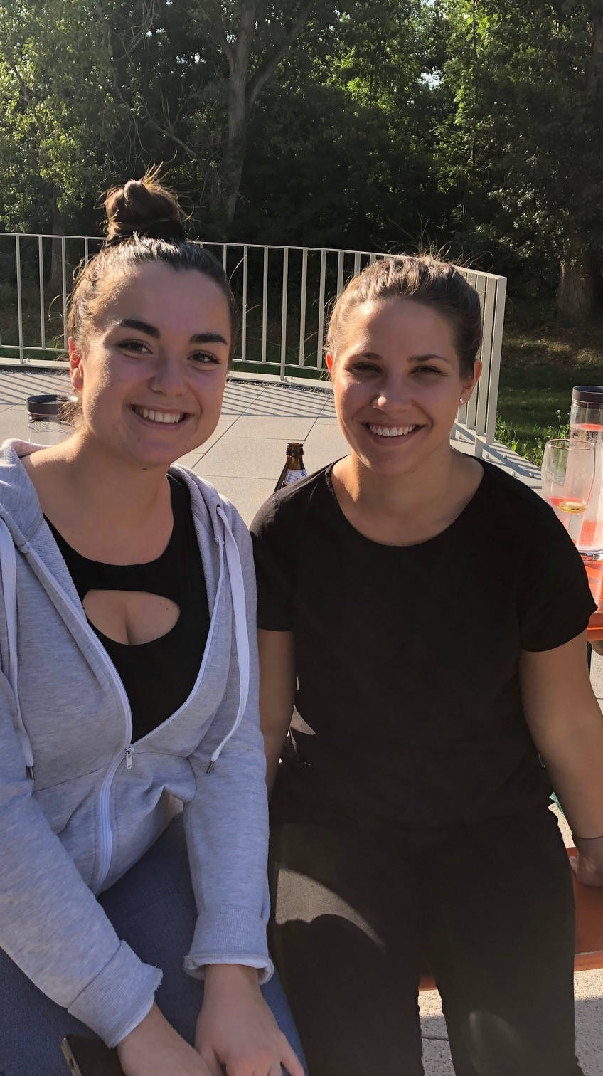 Zwei lächelnde Frauen sitzen an einem Tisch im Freien bei Sonnenschein, umgeben von Pflanzen.
