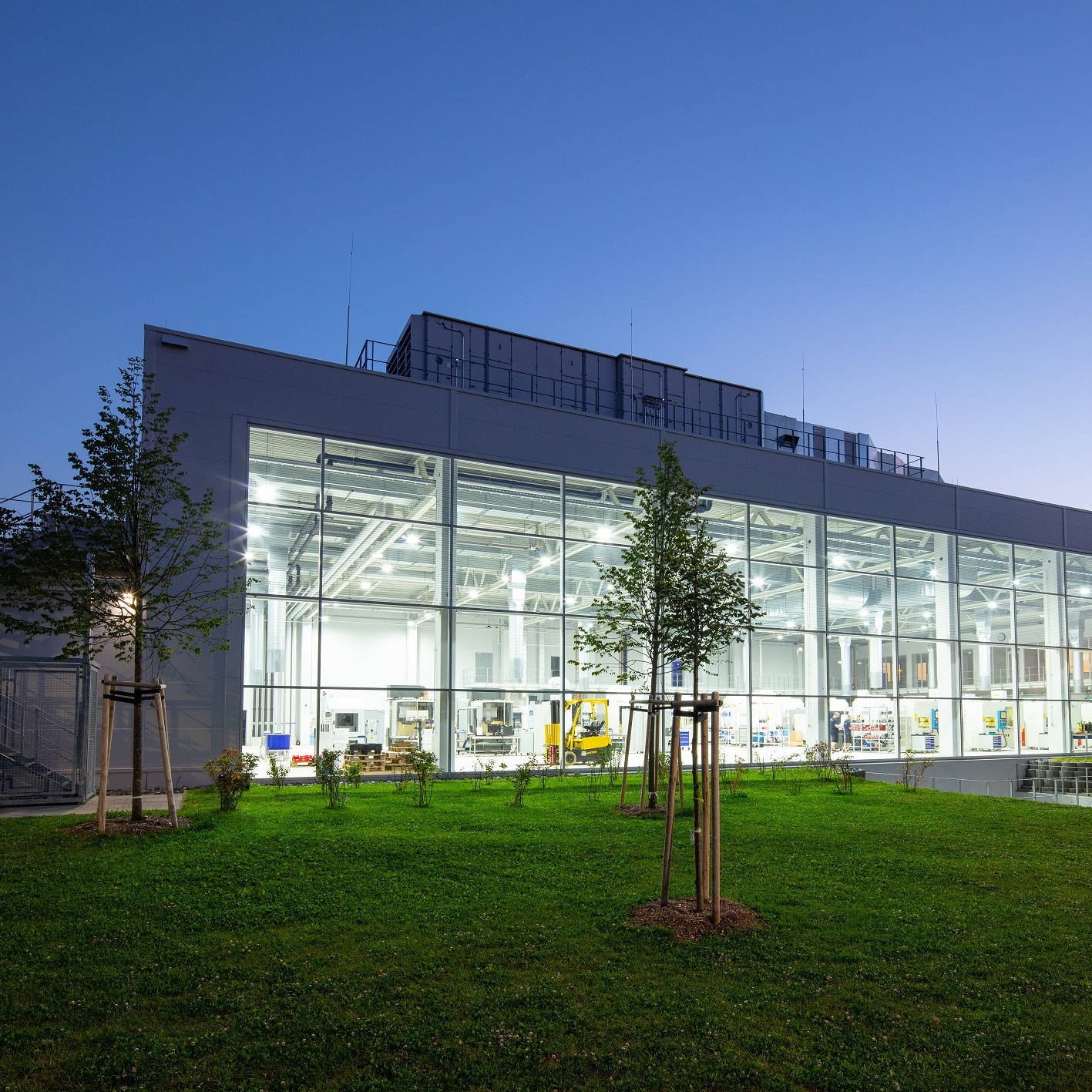 Moderne Fabrik mit Glasfassade bei Dämmerung, innen erleuchtet, umgeben von gepflegtem Rasen und jungen Bäumen.